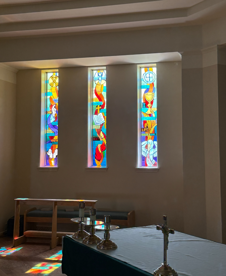 Stained glass windows depicting religious symbols and vibrant colors, enhancing a worship space at a church, with altar and candles in foreground.