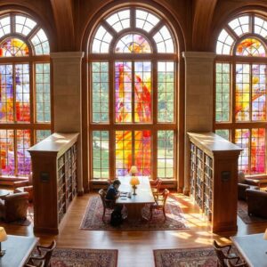 Library interior featuring vibrant stained glass windows, wooden bookshelves, and a study area with a person working at a table.