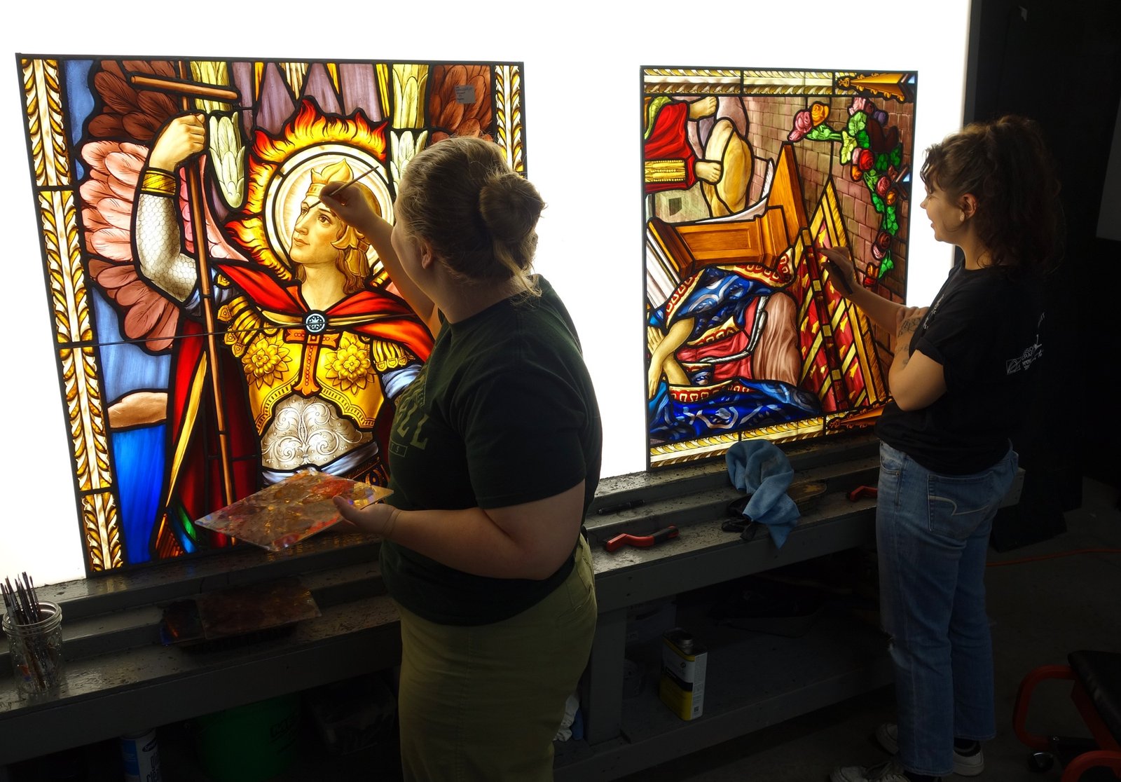 Two artisans painting intricate stained glass panels, showcasing detailed religious imagery and vibrant colors, in a workshop setting focused on restoration and preservation.