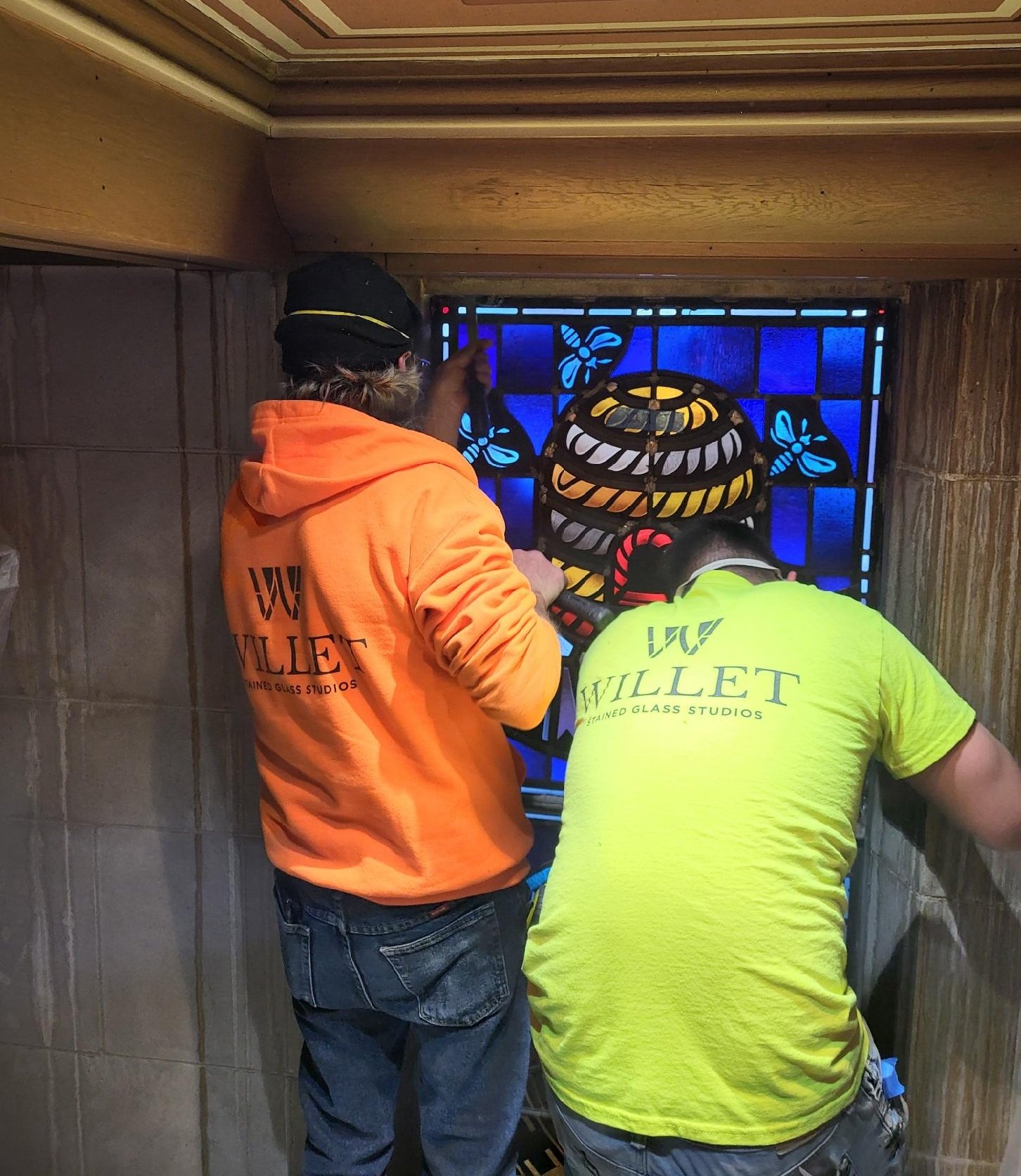 Two Willet Studios employees working on stained glass installation, featuring a colorful window design with bees and decorative elements, highlighting craftsmanship and collaboration in art preservation.