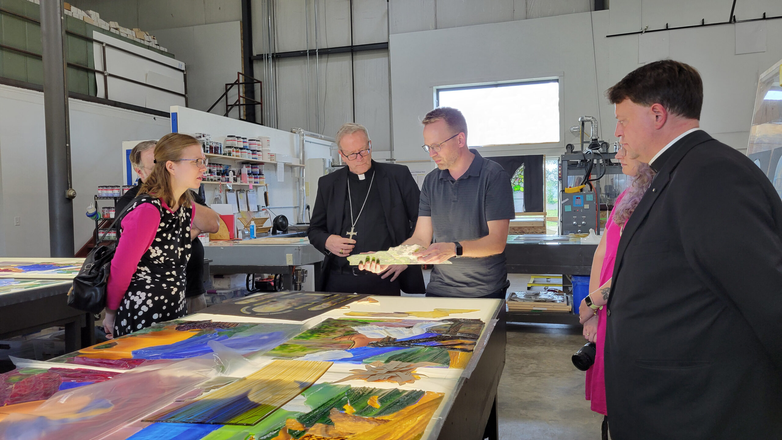 Group of individuals, including clergy, observing stained glass designs and discussing artistic elements in a workshop setting at Willet Stained Glass Studios.