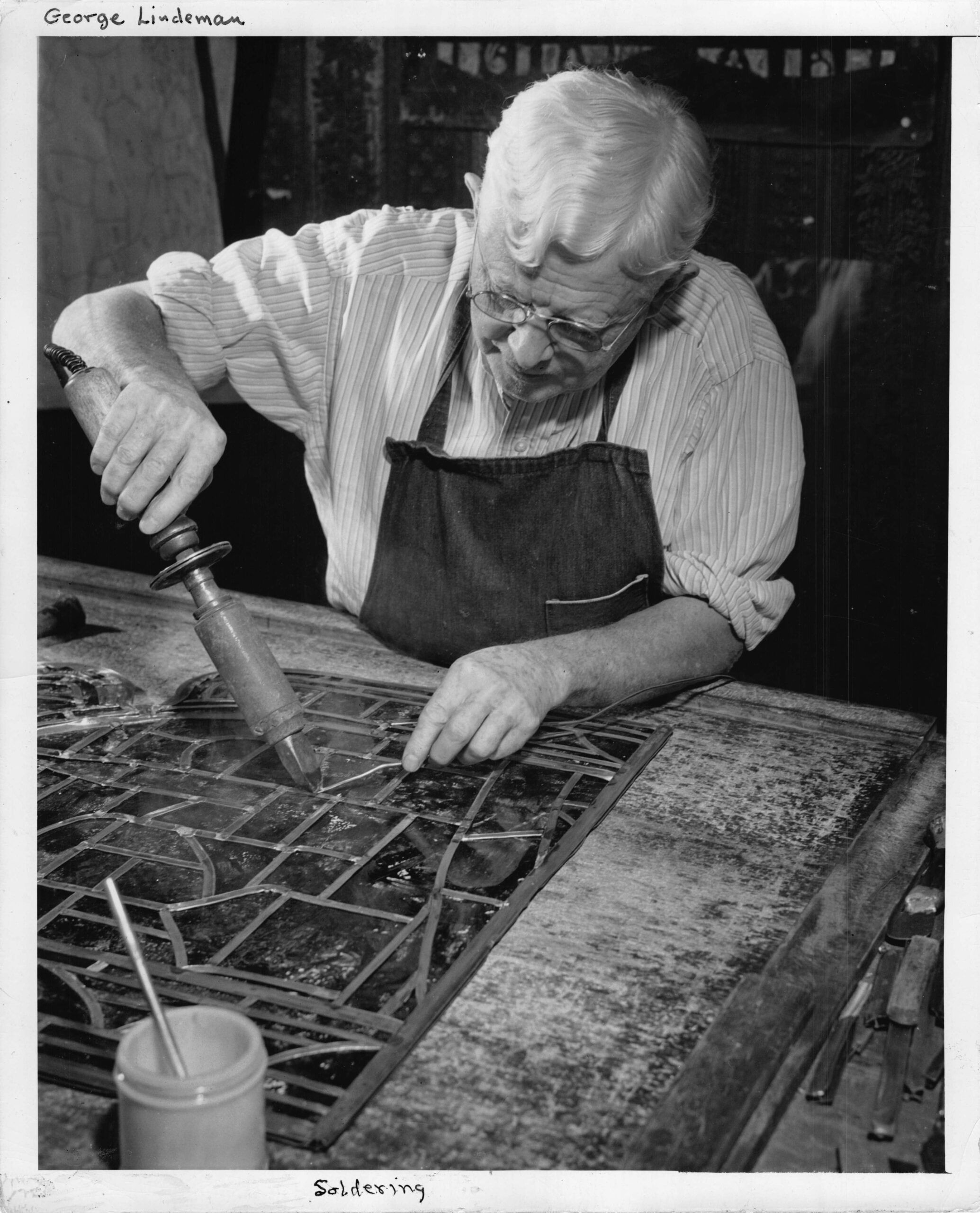George Lindeman soldering stained glass, demonstrating craftsmanship in stained glass artistry, with tools and materials visible on the workbench, reflecting the legacy of Willet Stained Glass Studios.