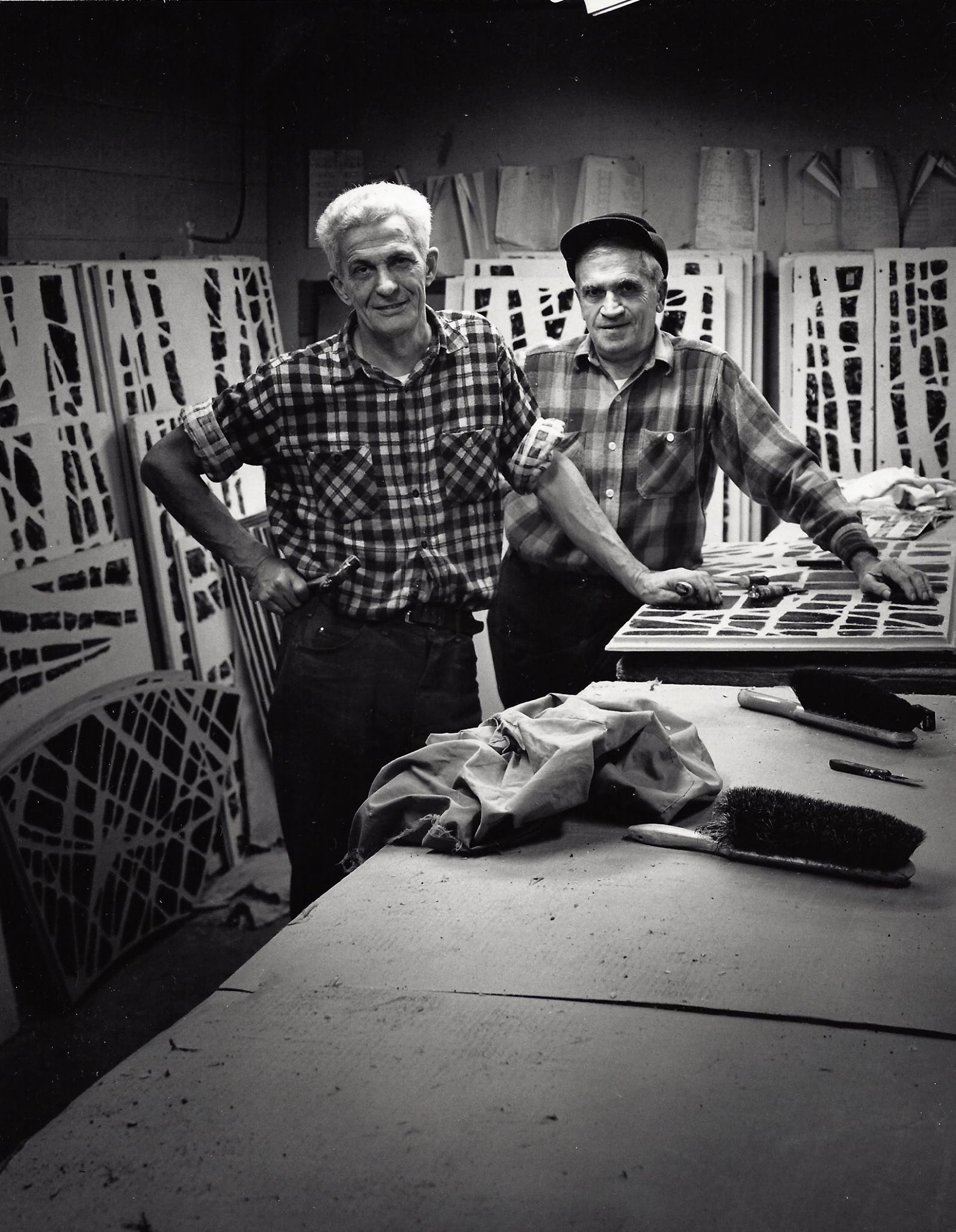 Two stained glass artisans in a workshop, surrounded by glass panels and tools, showcasing craftsmanship at Willet Stained Glass Studios.