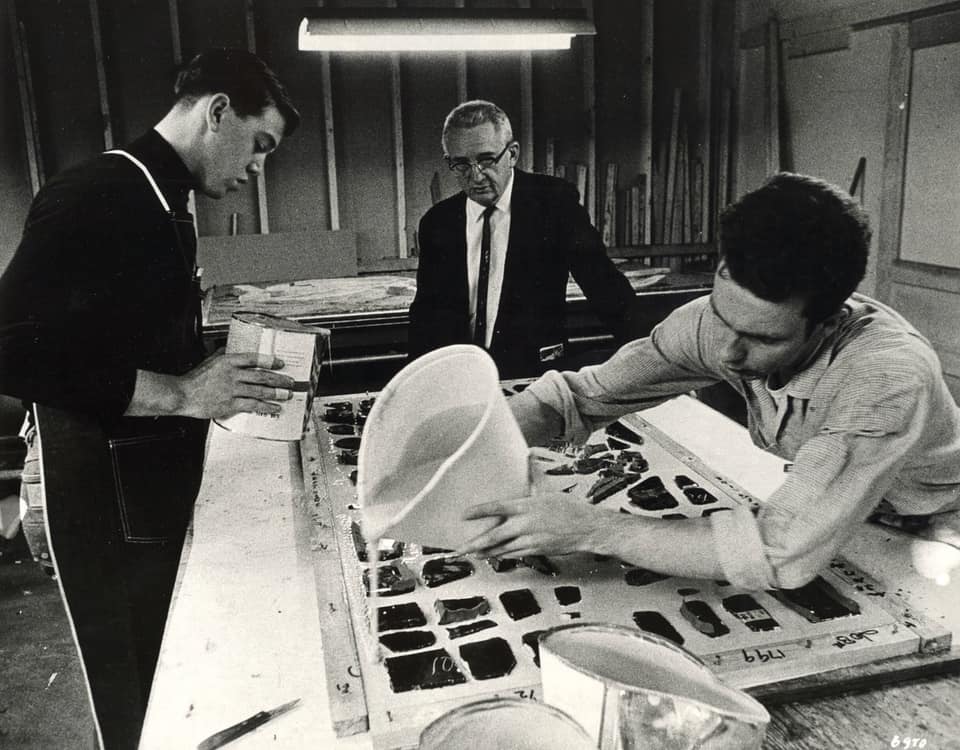 Three artisans collaboratively working on stained glass artwork, with one pouring liquid into a mold, another holding a container, and a third observing, reflecting the craftsmanship and design excellence of Willet Stained Glass Studios.