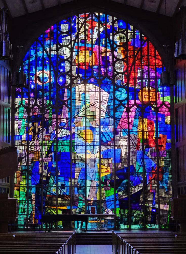 Stained glass window featuring vibrant colors and abstract designs, showcasing artistry by Willet Stained Glass Studios, set in a sacred space with a piano and pews.