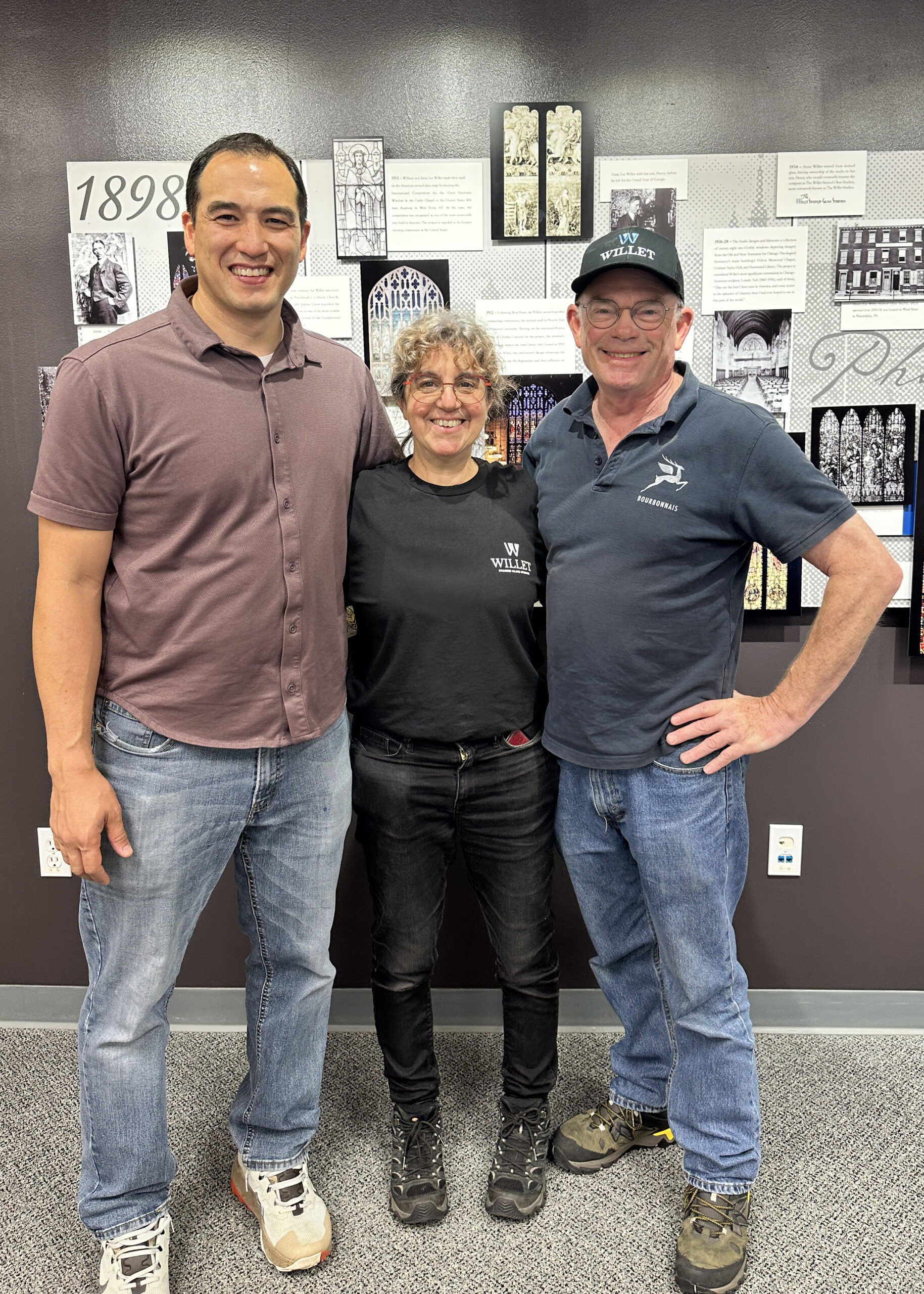 Three individuals posing together in front of a wall featuring stained glass designs and historical information about Willet Stained Glass Studios, highlighting the studio's legacy and craftsmanship in stained glass artistry.