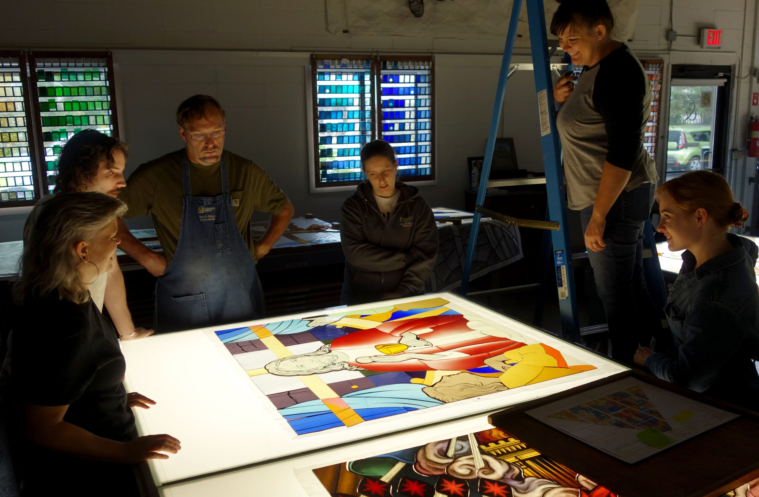 Group of stained glass artists collaborating around a light table, reviewing a colorful stained glass design, with vibrant glass samples visible in windows, emphasizing teamwork and artistic process at Willet Studios.
