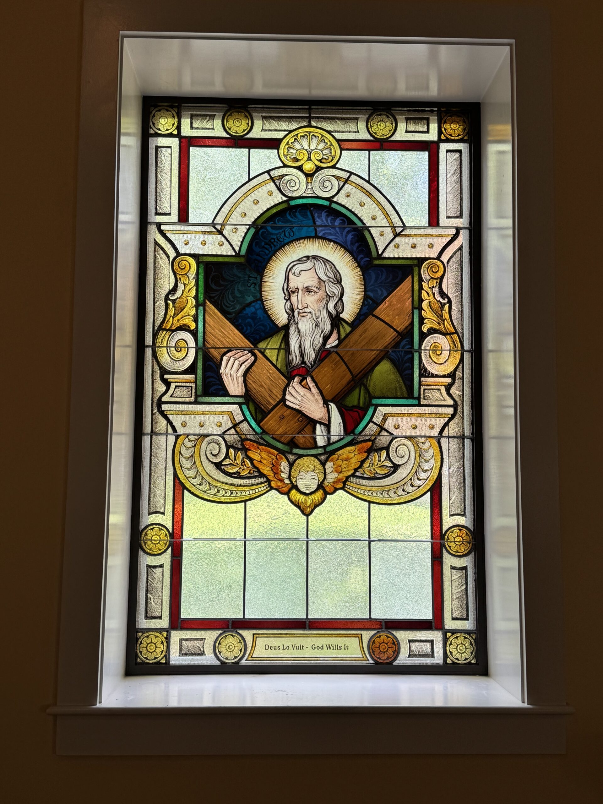 Stained glass window featuring a bearded figure holding wooden beams, surrounded by ornate designs and the inscription "Deus Lo Vult - God Wills It," symbolizing faith and inspiration in a sacred space.