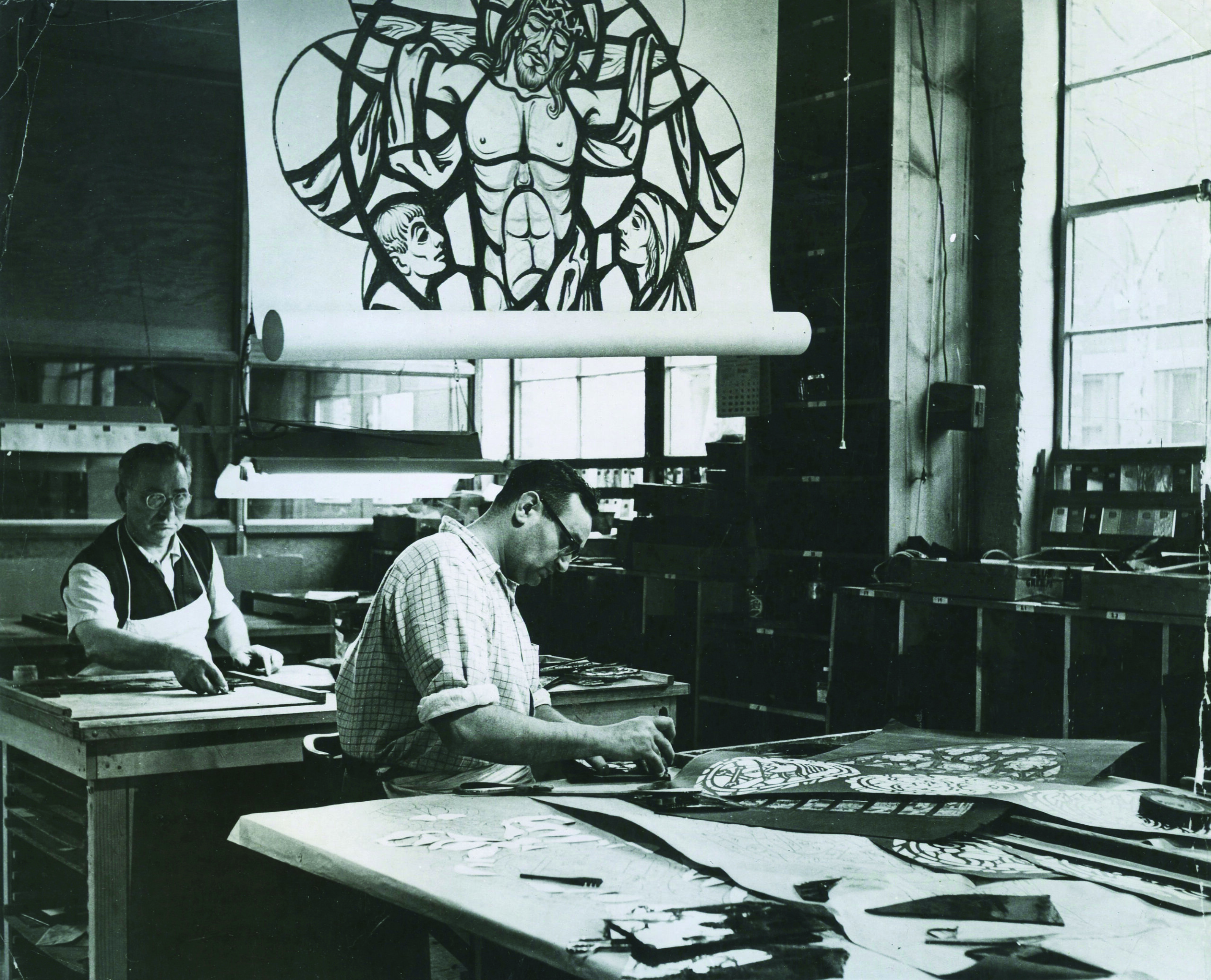 Two artisans working on stained glass designs in a studio, featuring a large stained glass pattern of a religious figure above, with tools and glass pieces on the table, highlighting craftsmanship and artistry in stained glass production.