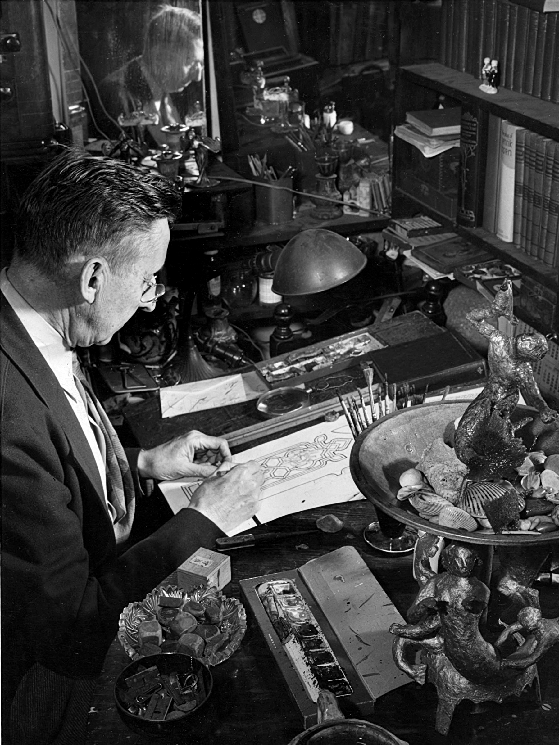 Artist sketching stained glass design at cluttered workspace, featuring tools, paints, and decorative objects, reflecting Willet Stained Glass Studios' craftsmanship and artistry legacy.