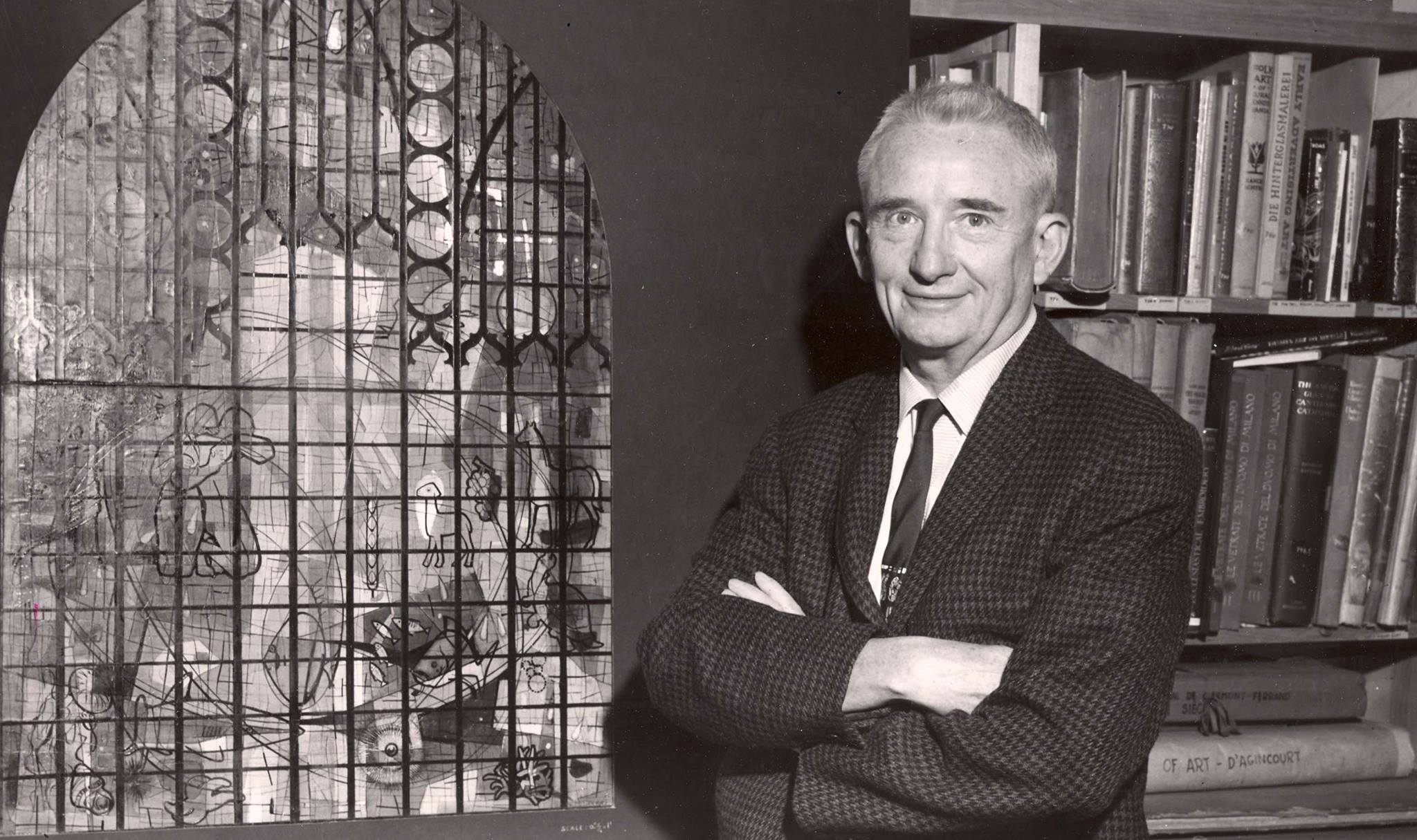 Henry Lee Willet standing beside a stained glass design, showcasing artistry and craftsmanship in stained glass, with bookshelves in the background reflecting the studio's historical legacy.