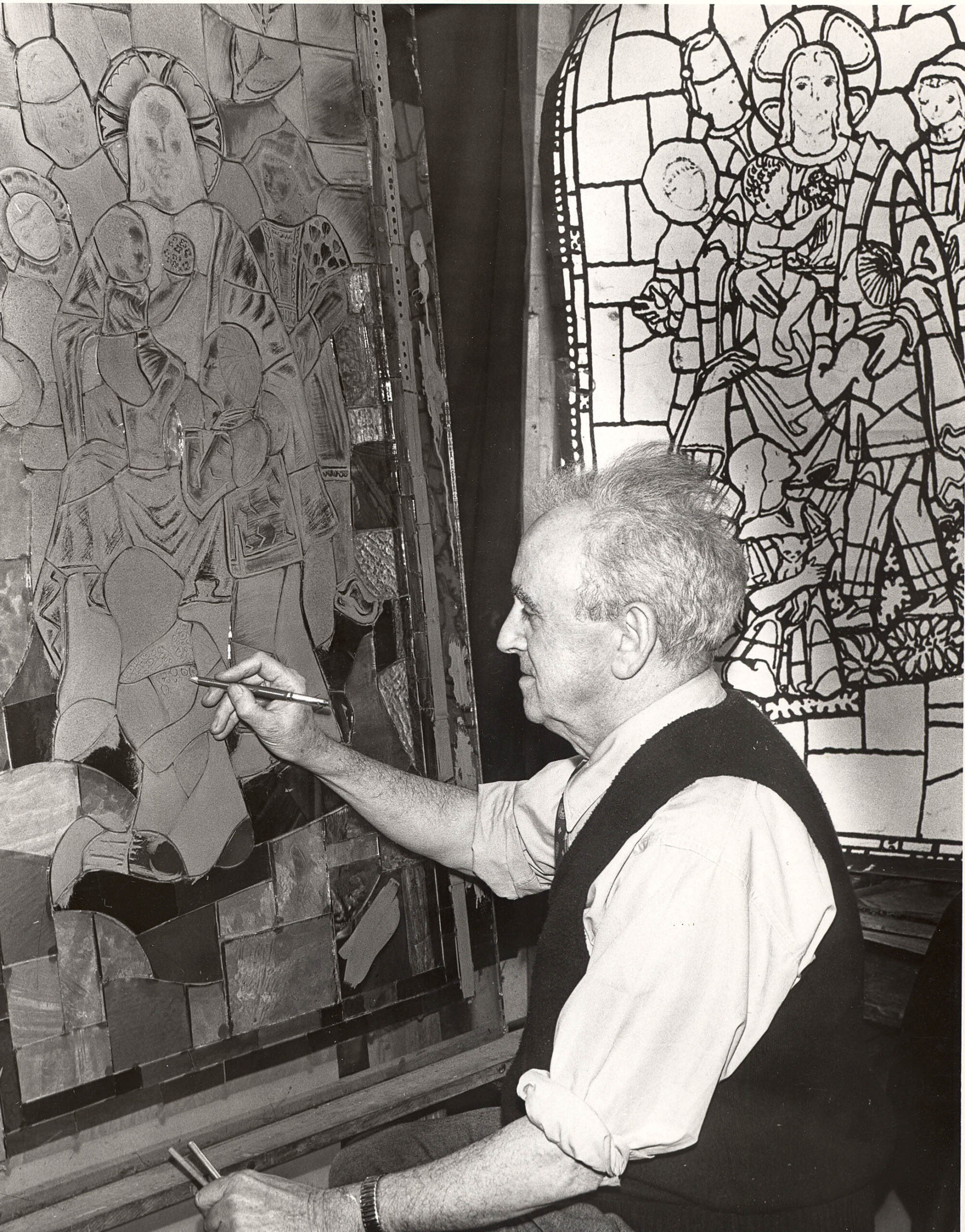 Artist painting stained glass panel, showcasing craftsmanship and artistry in stained glass design, with a completed window in the background, reflecting Willet Stained Glass Studios' legacy.