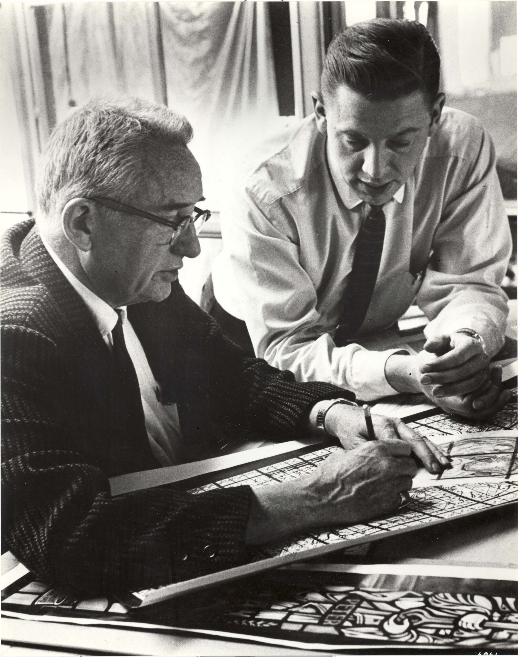 Two men collaborating on stained glass design, one sketching on a panel while the other observes, showcasing craftsmanship and artistry at Willet Stained Glass Studios.