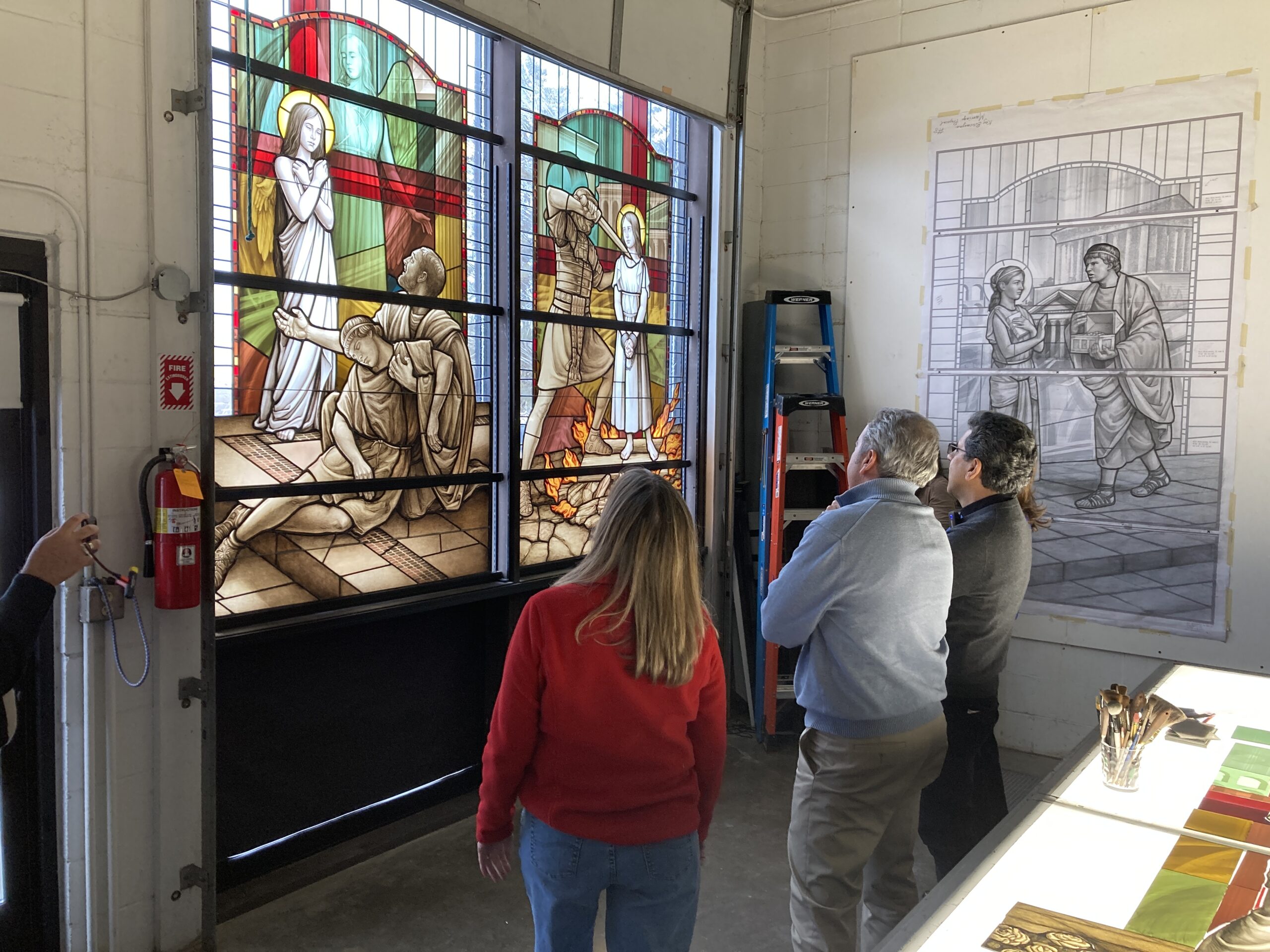 Group of people observing a colorful stained glass window design depicting biblical scenes, with a sketch of another stained glass window in the background, highlighting the collaborative process of creating religious art.