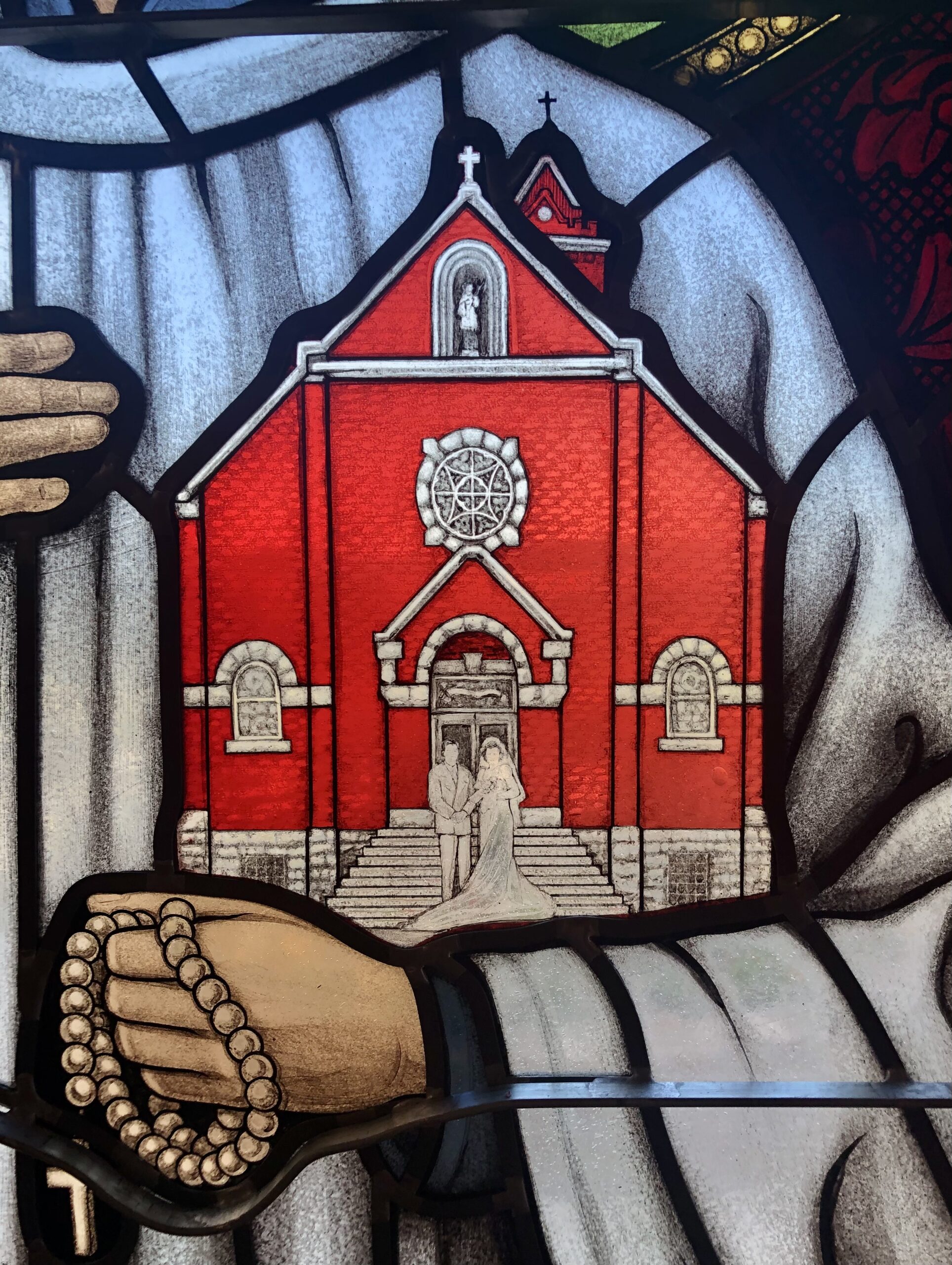 Stained glass depiction of a red church with a couple in wedding attire, framed by a hand holding rosary beads, illustrating the integration of art and faith in religious spaces.