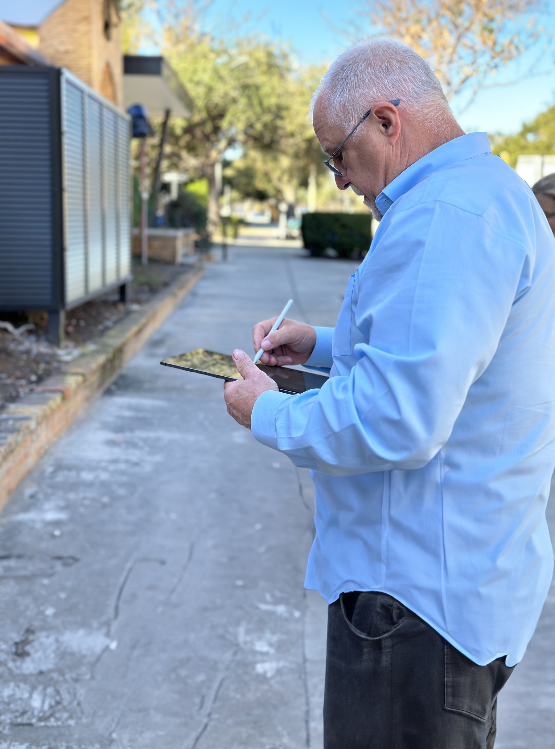 Man in blue shirt using a tablet and stylus while conducting an on-site assessment for stained glass consulting, with a sidewalk and greenery in the background.