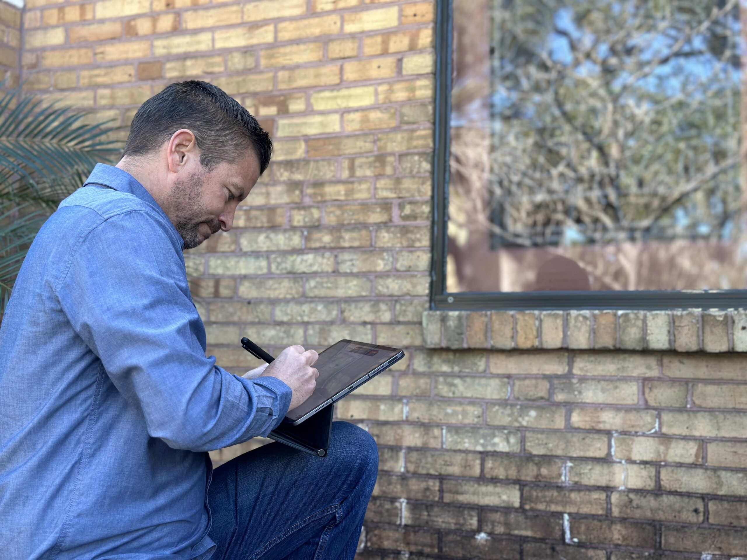 Man inspecting and taking notes on a tablet while seated next to a brick wall, with a window visible in the background, related to stained glass consulting services at Willet Studios.