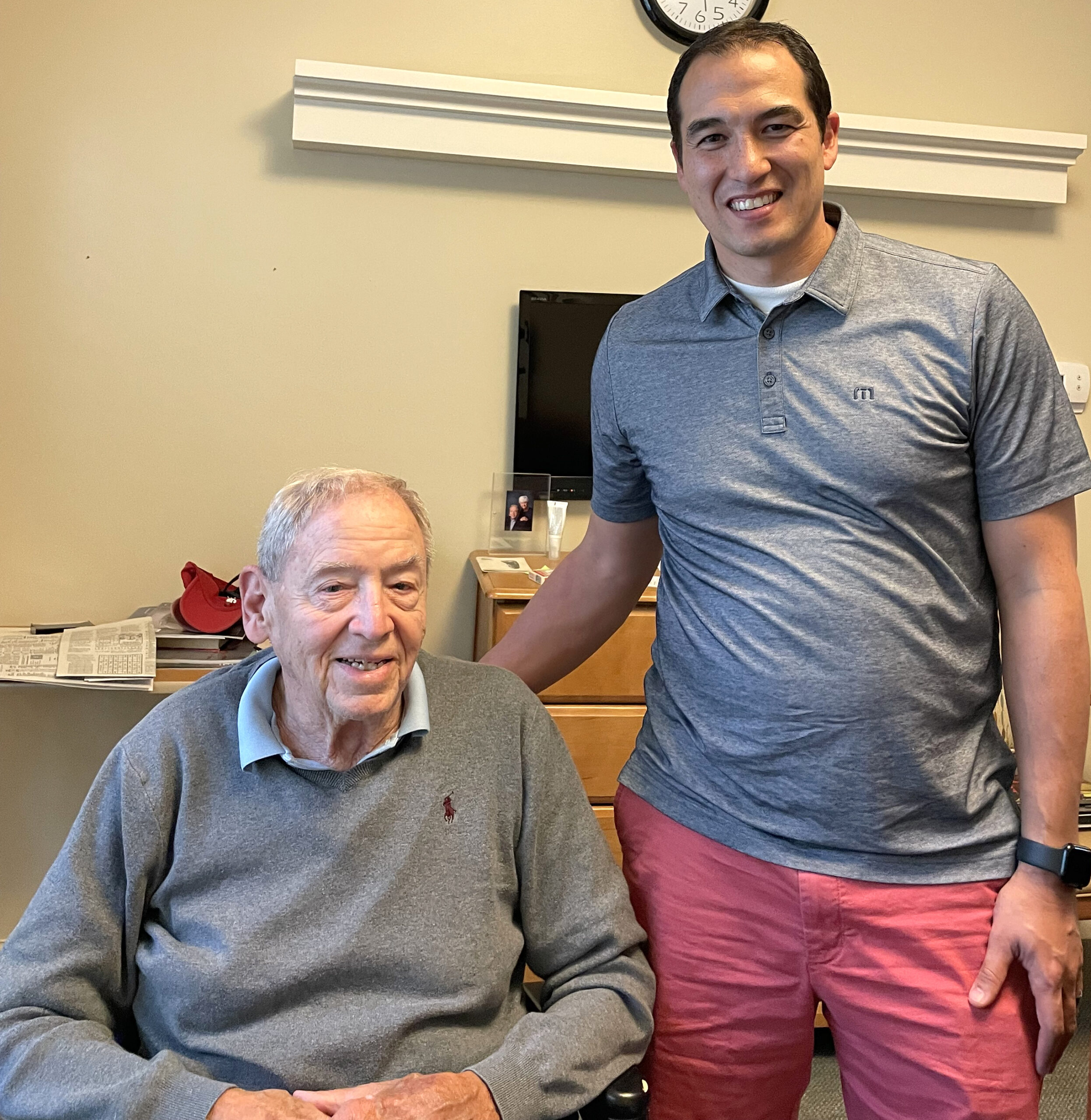 Elderly man smiling in a wheelchair with younger man standing beside him, in a cozy indoor setting, reflecting themes of legacy and connection.