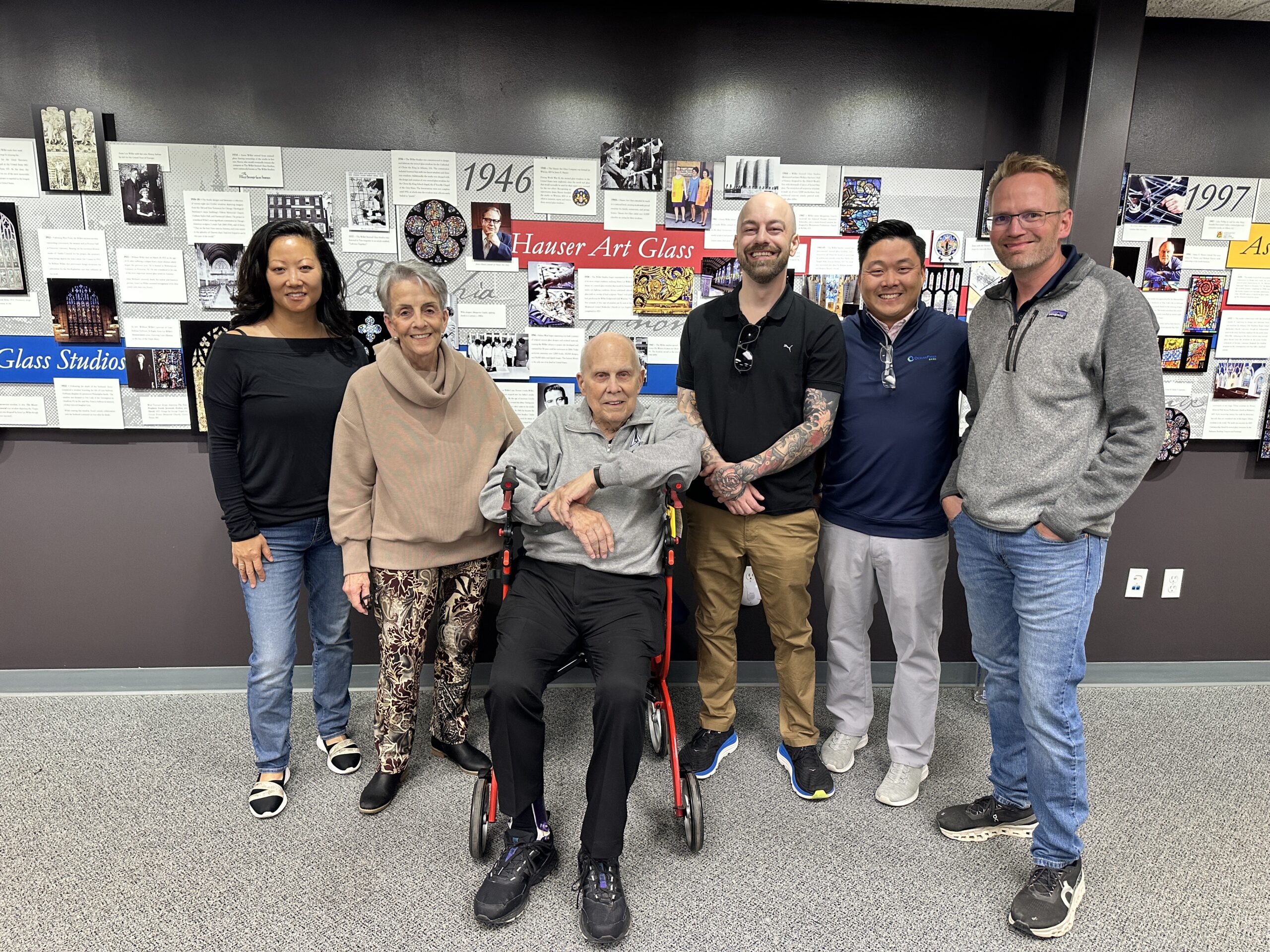 Group of six individuals posing in front of a historical display about Willet Stained Glass Studios, showcasing their legacy and milestones in stained glass artistry.