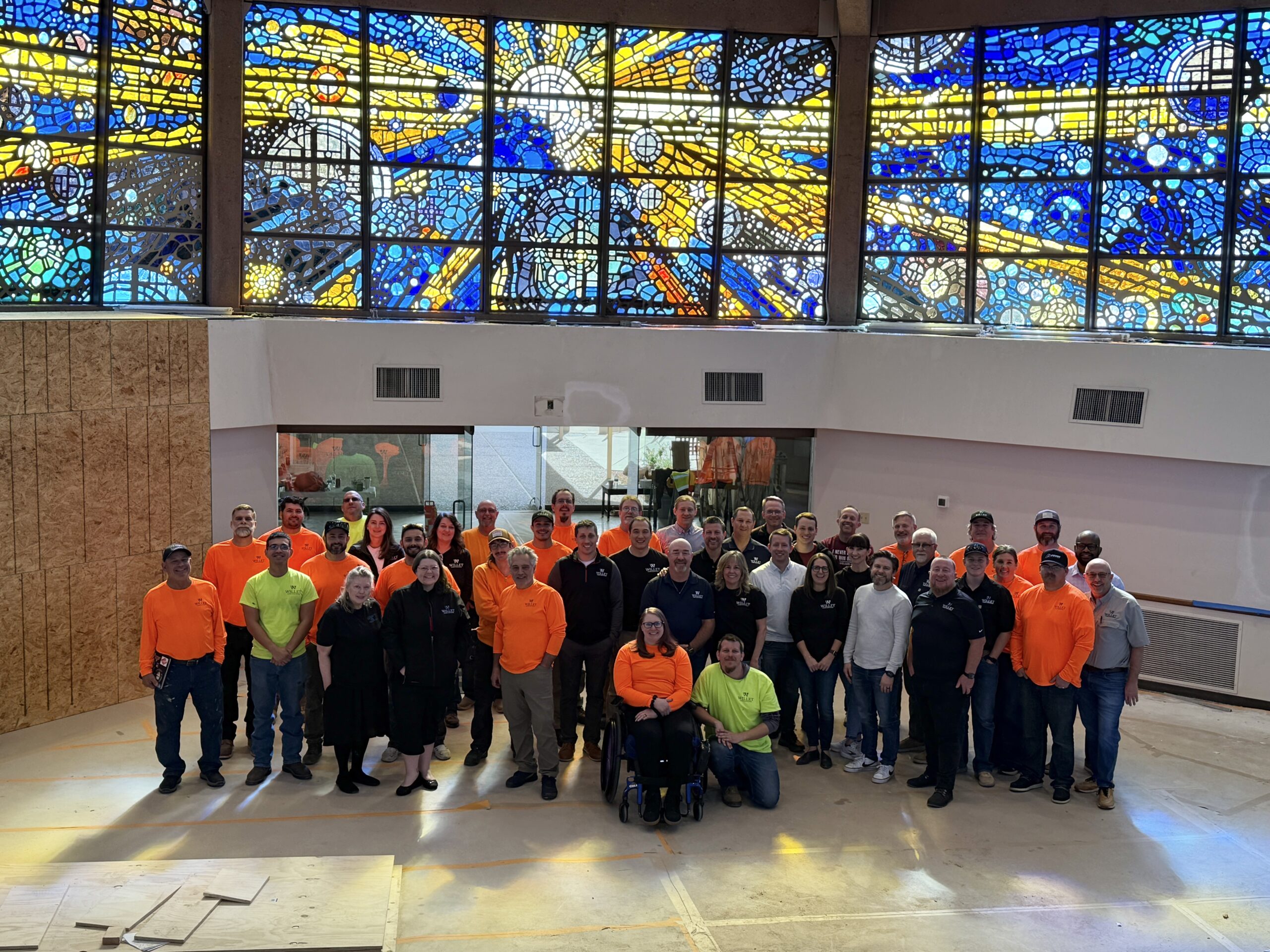 Group of Willet Stained Glass Studios team members in bright work attire, gathered in front of a vibrant stained glass window, showcasing collaboration and craftsmanship in an architectural space.