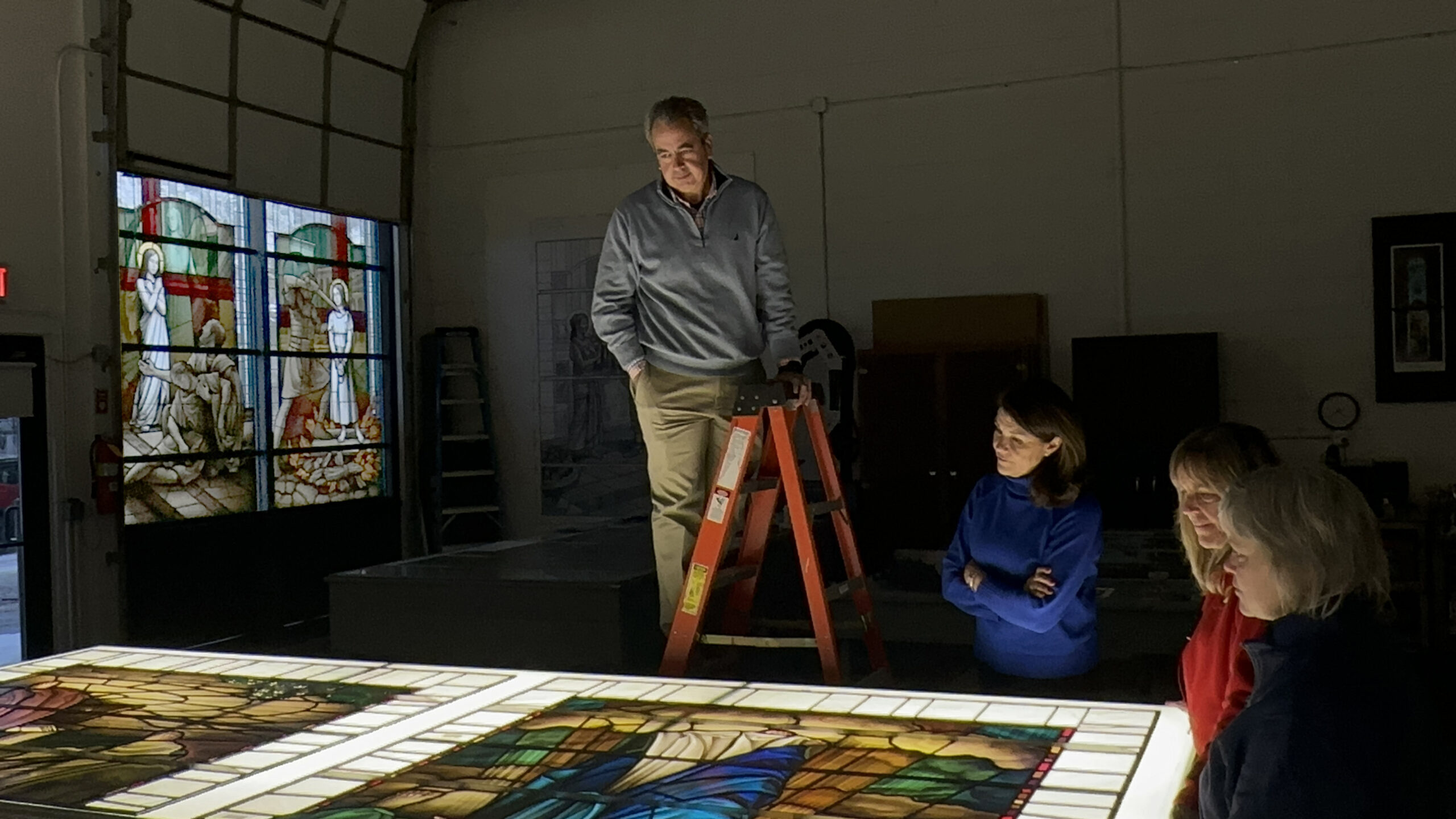 Professional overseeing stained glass evaluation with team, illuminated glass panels, workshop setting, focus on restoration and preservation.