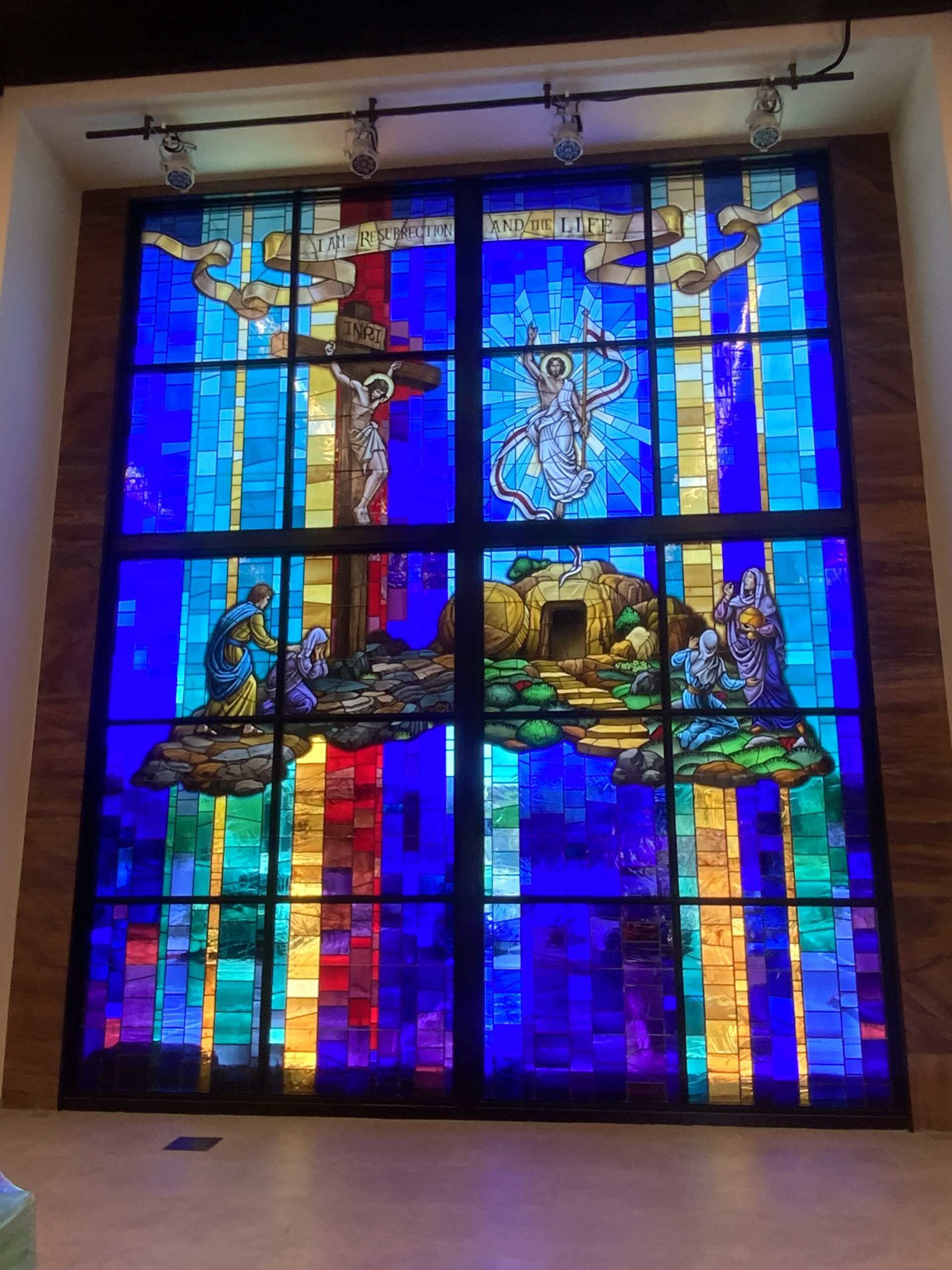 Stained glass window depicting the resurrection of Christ, featuring vibrant blue and gold hues, biblical figures, and the inscription "I am the resurrection and the life," showcasing Willet Stained Glass Studios' artistry in religious spaces.