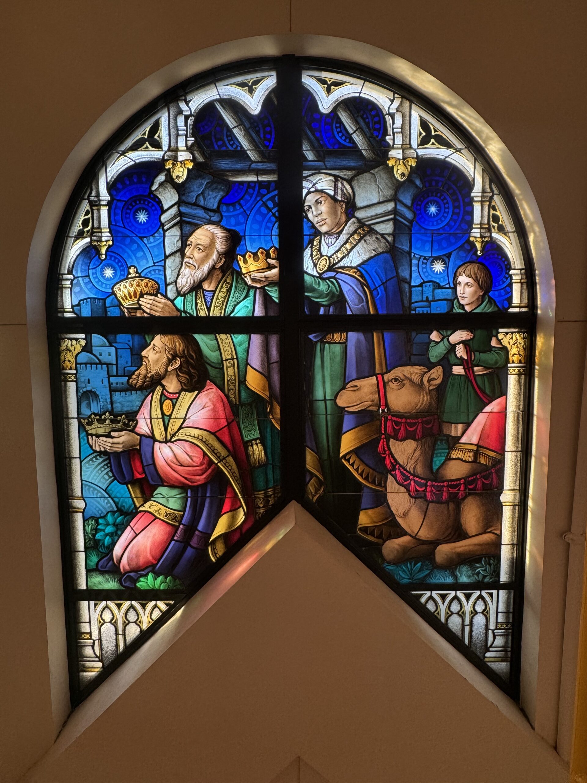 Stained glass window depicting three wise men presenting gifts, a woman holding a crown, and a young boy with a camel, showcasing vibrant colors and intricate details, symbolizing faith and worship in a religious setting.