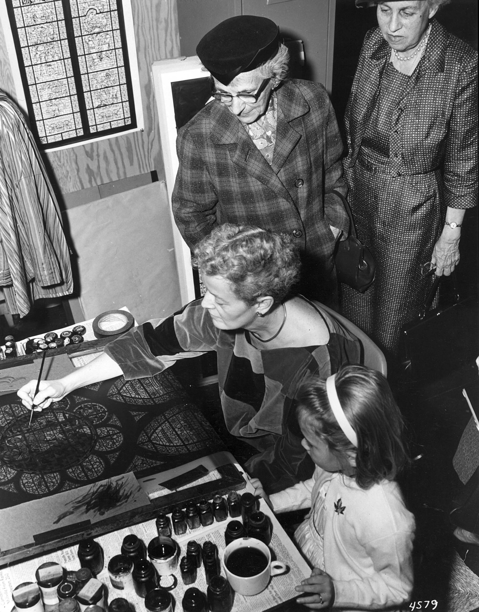Woman painting stained glass design with child observing, surrounded by paint bottles and art materials, showcasing craftsmanship and artistry in stained glass, reflecting Willet Stained Glass Studios' legacy.