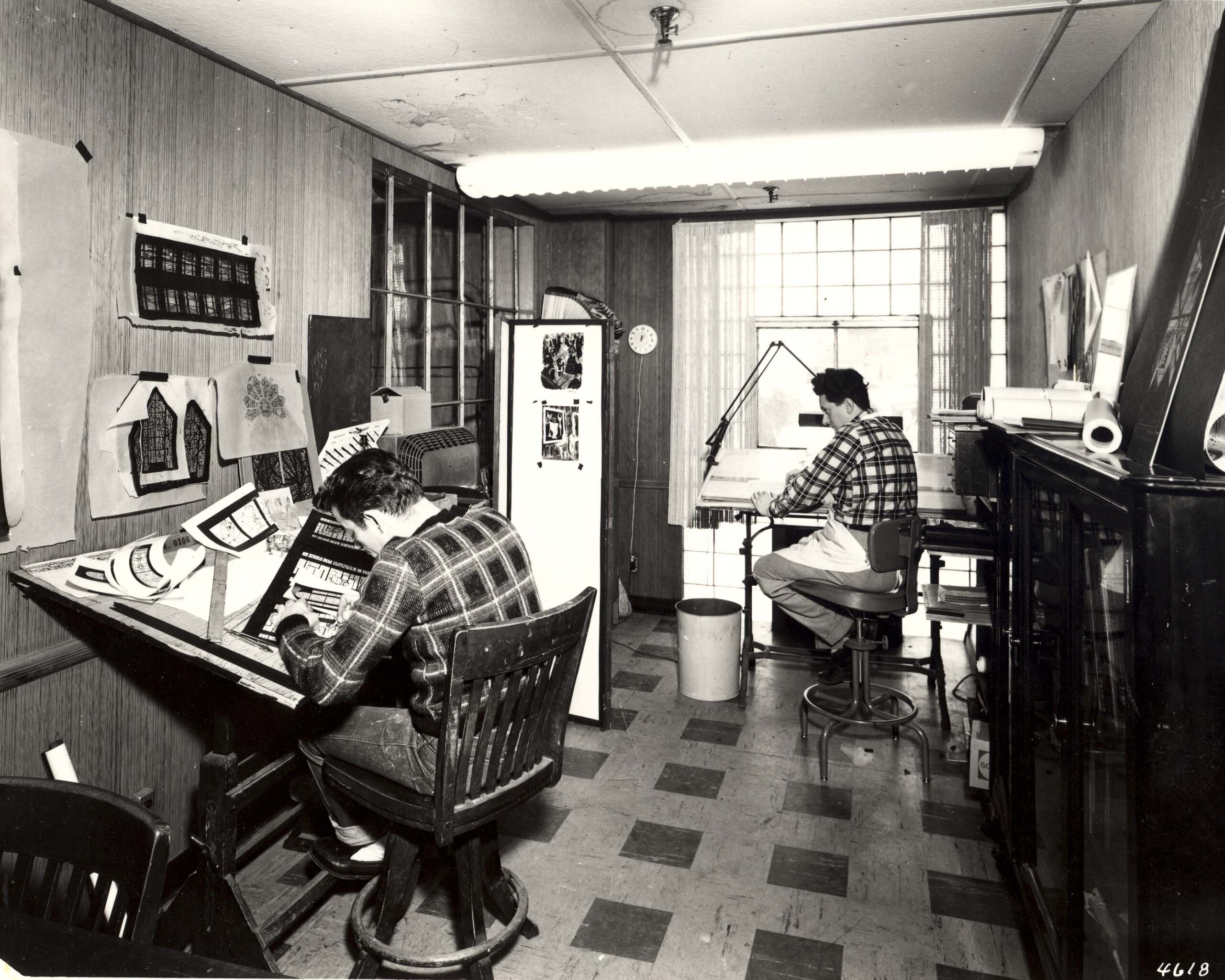 Artists working in stained glass studio, one sketching designs at a drafting table, the other at a drawing board, surrounded by stained glass patterns and tools, reflecting craftsmanship and artistry at Willet Stained Glass Studios.