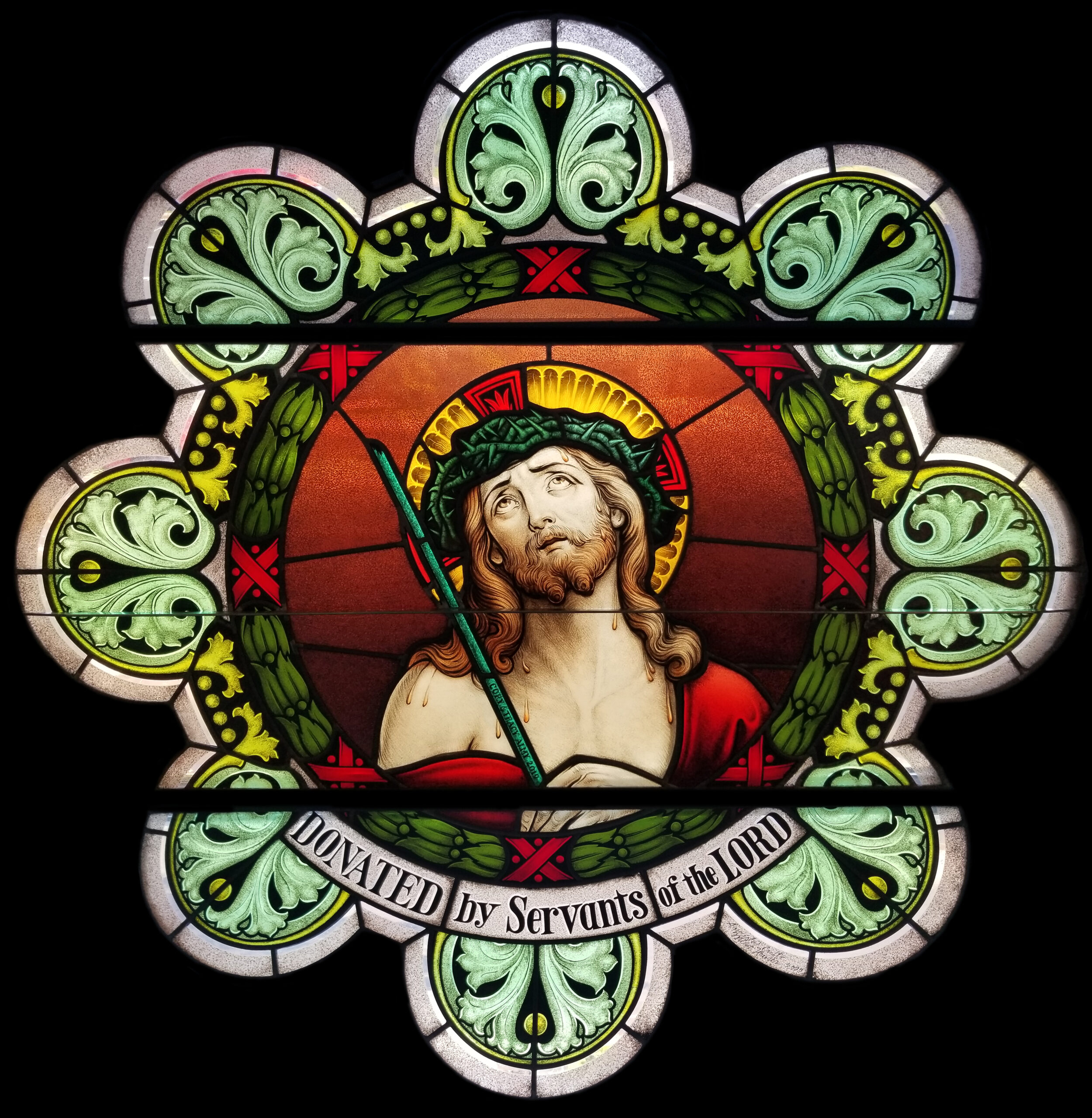 Stained glass window depicting Jesus Christ with a crown of thorns and a staff, framed by intricate green foliage and a banner reading "DONATED by Servants of the LORD," symbolizing faith and dedication in worship spaces.