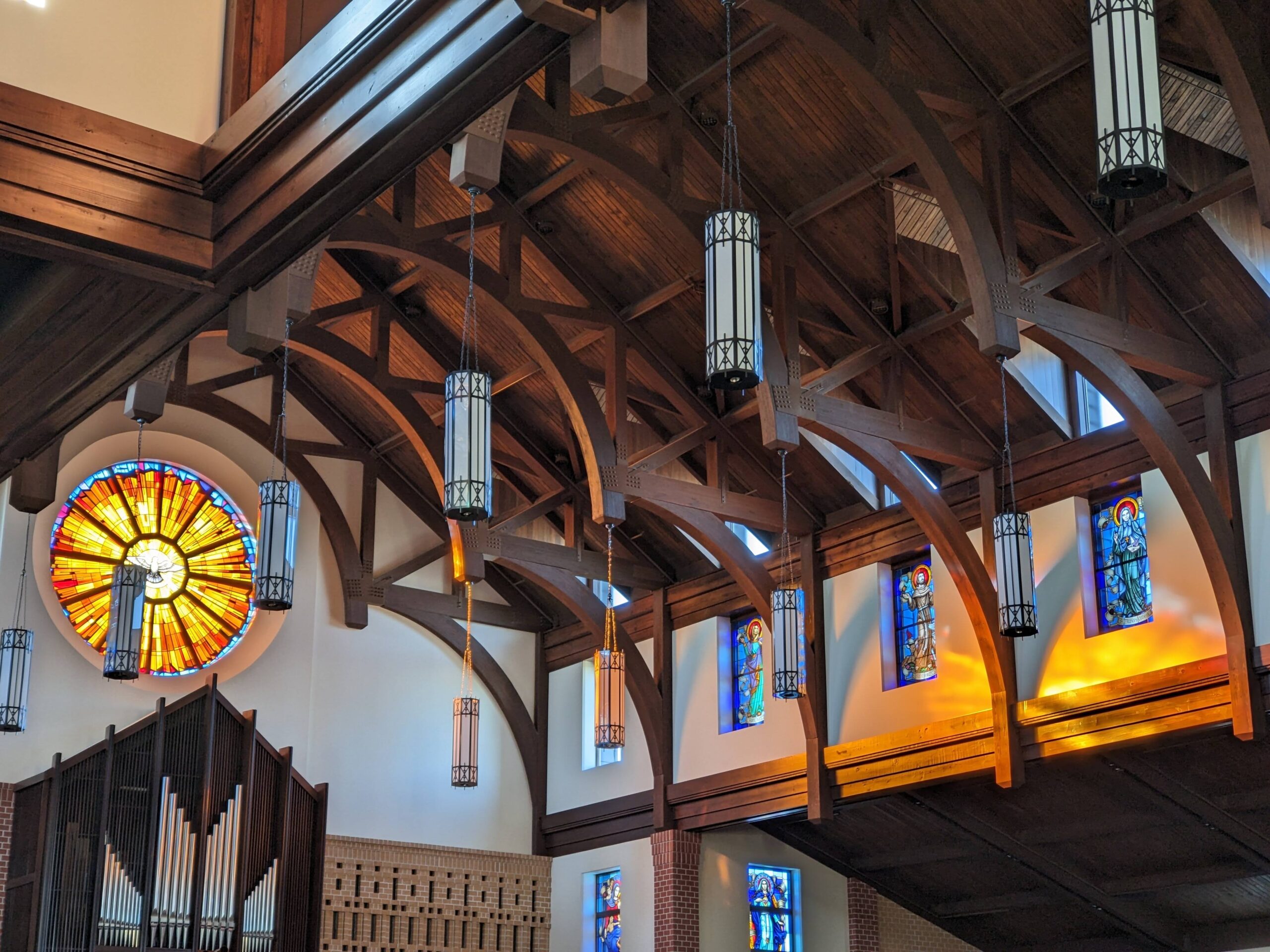 Interior of a church featuring vibrant stained glass windows, including a large circular rose window with warm colors, decorative hanging light fixtures, and wooden beams, showcasing the artistry of Willet Stained Glass Studios.