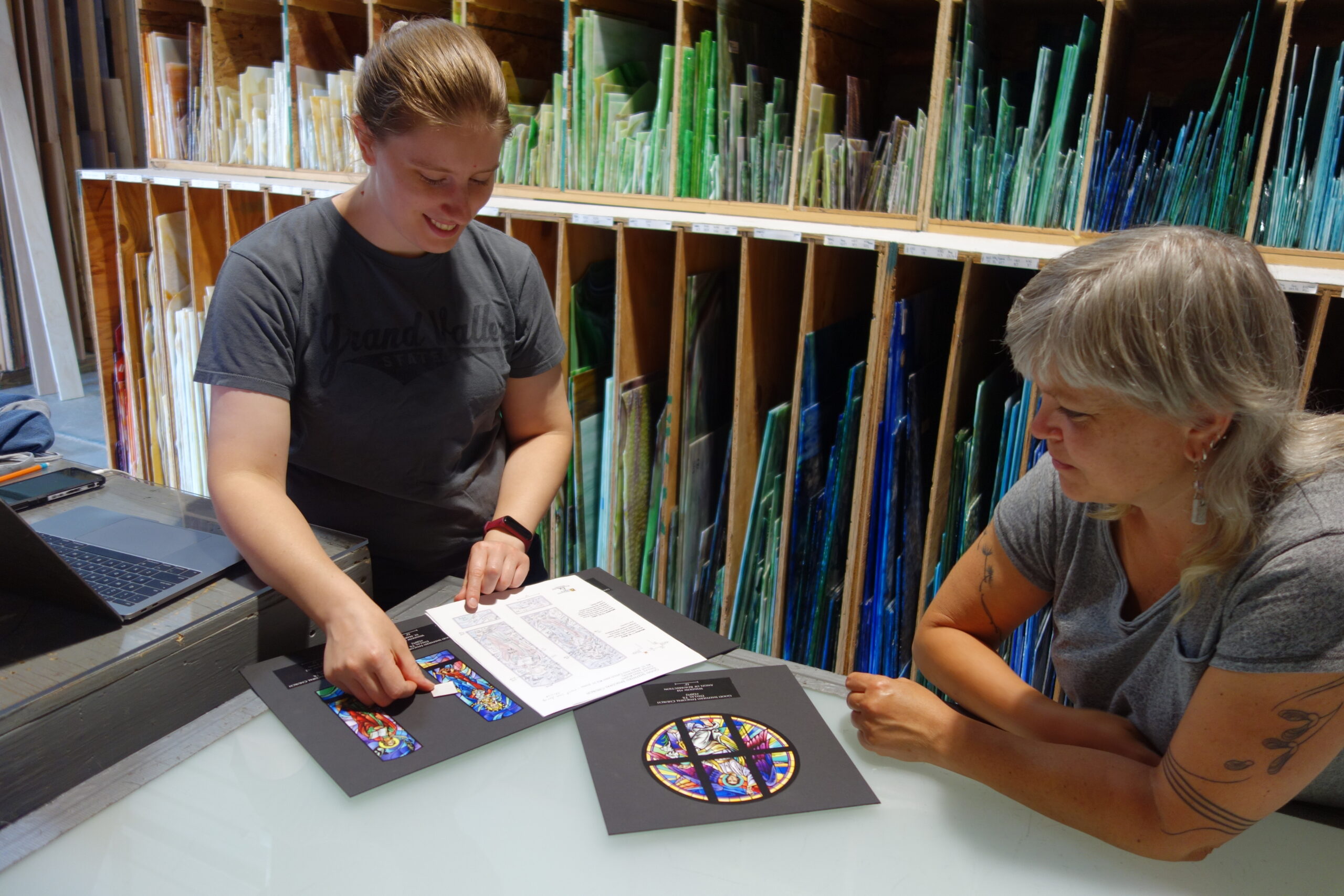 Two stained glass artists collaborating at a table, reviewing design sketches and glass samples, surrounded by colorful glass panels.