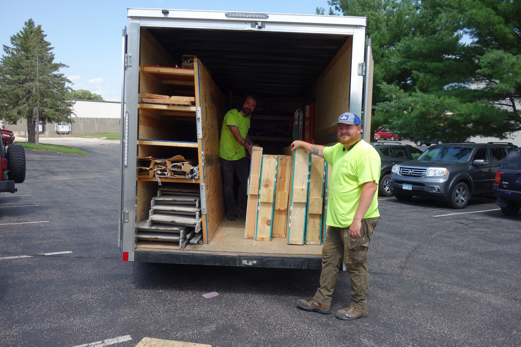 Two Willet Studios team members in bright yellow shirts unloading materials from a trailer, preparing for stained glass window installation at a project site.