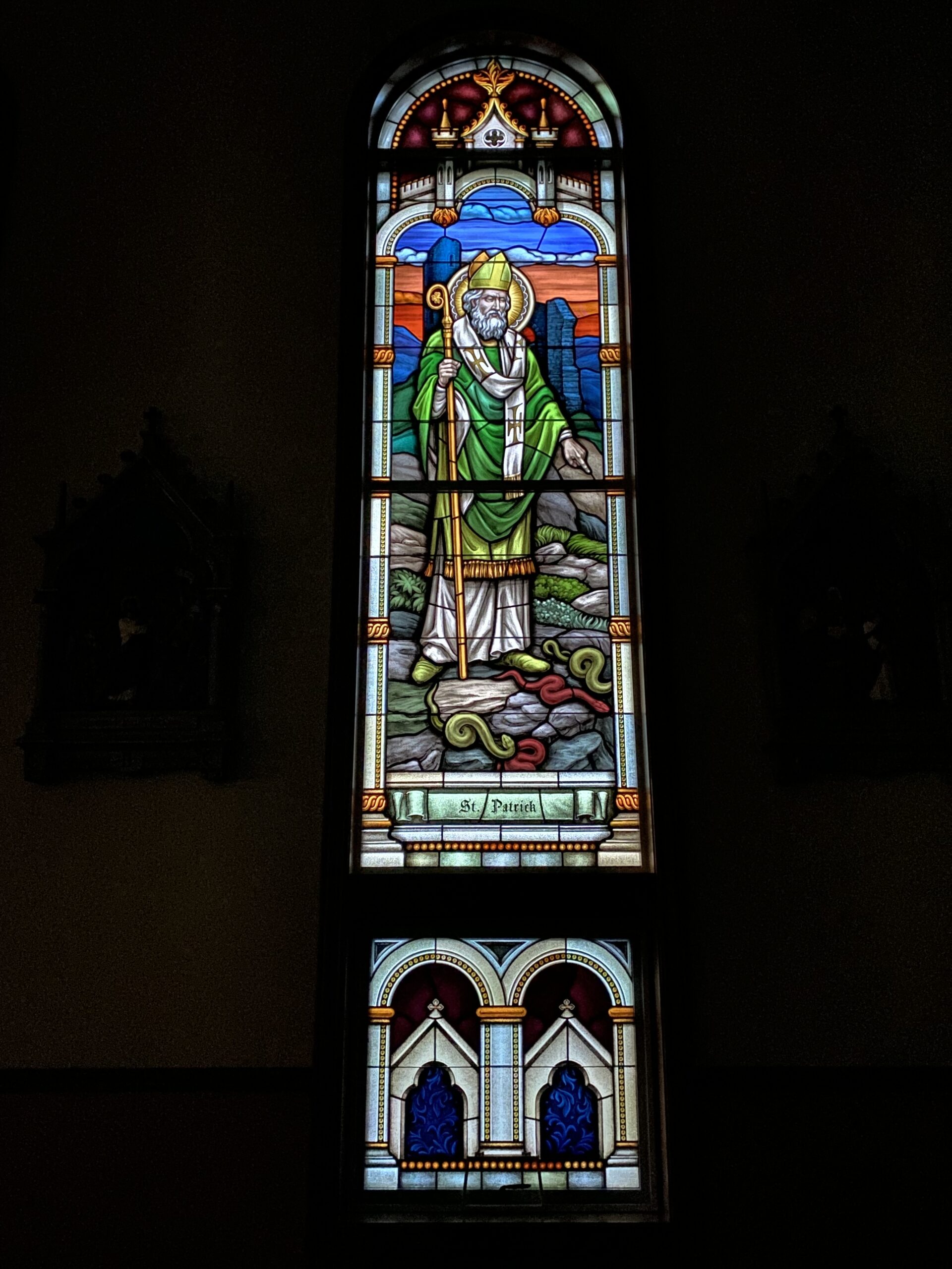 Stained glass window depicting St. Patrick in a green robe with a staff, surrounded by mountains and serpents, showcasing Willet Stained Glass Studios' artistry for worship spaces.