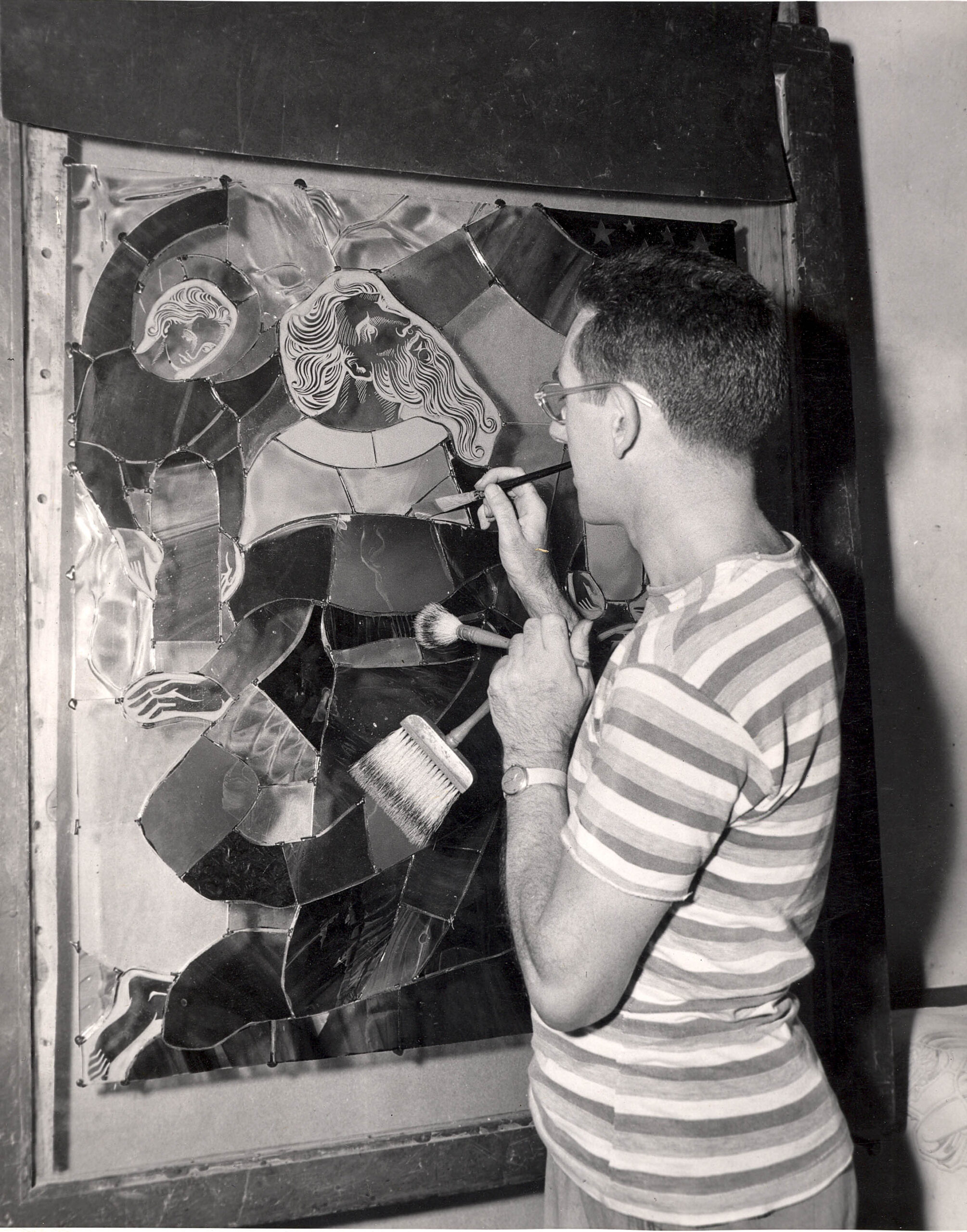 Artist working on stained glass panel, featuring figures and intricate designs, showcasing craftsmanship in stained glass artistry at Willet Stained Glass Studios.