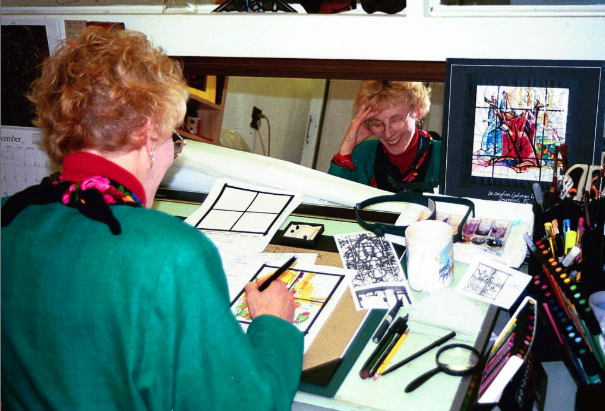 Artist working on stained glass design at a drafting table, surrounded by sketches, tools, and colorful materials, reflecting Willet Stained Glass Studios' commitment to artistry and craftsmanship.