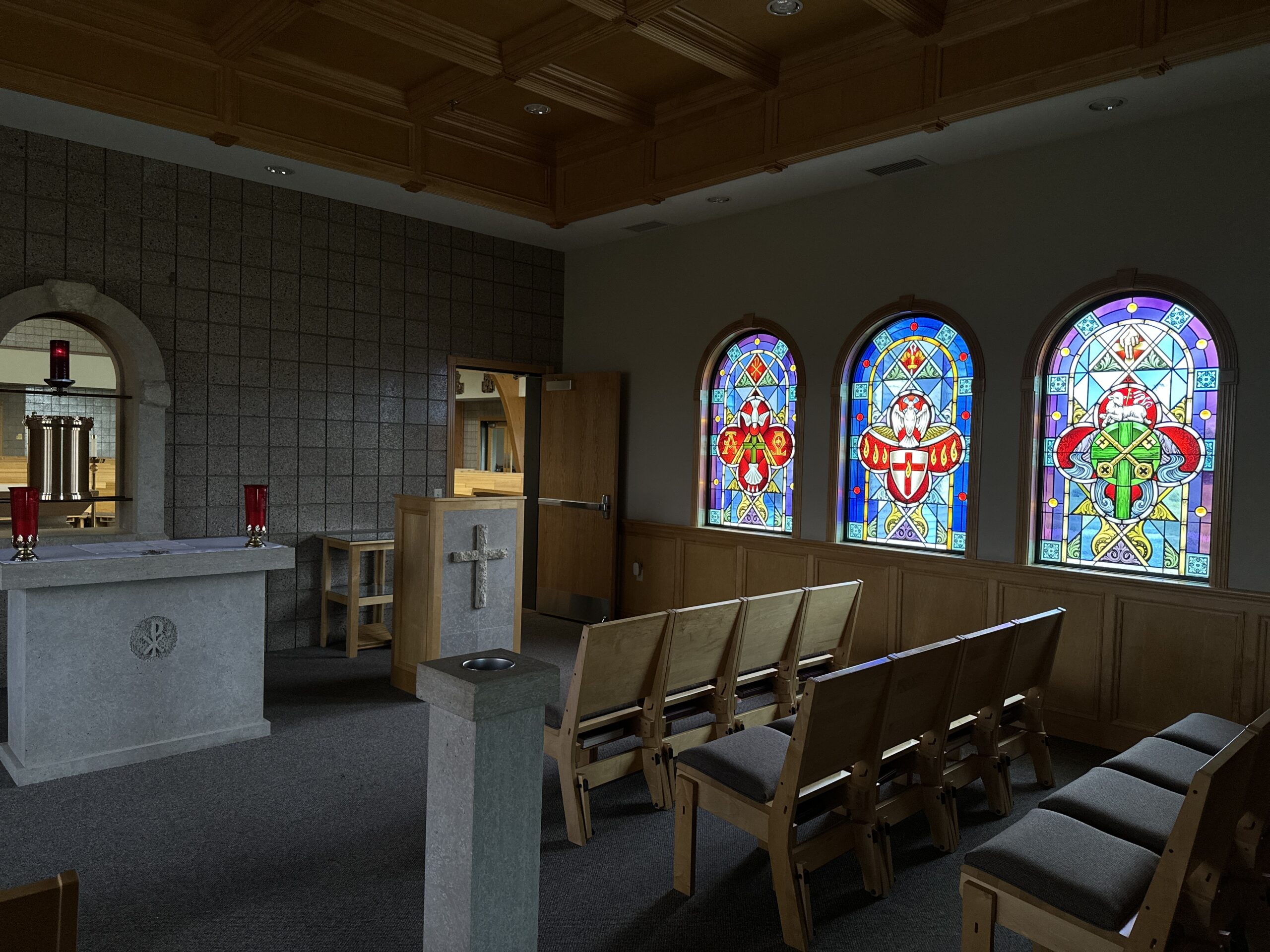 Interior of a chapel featuring three vibrant stained glass windows depicting religious symbols, an altar with candles, and wooden seating arranged for worship.