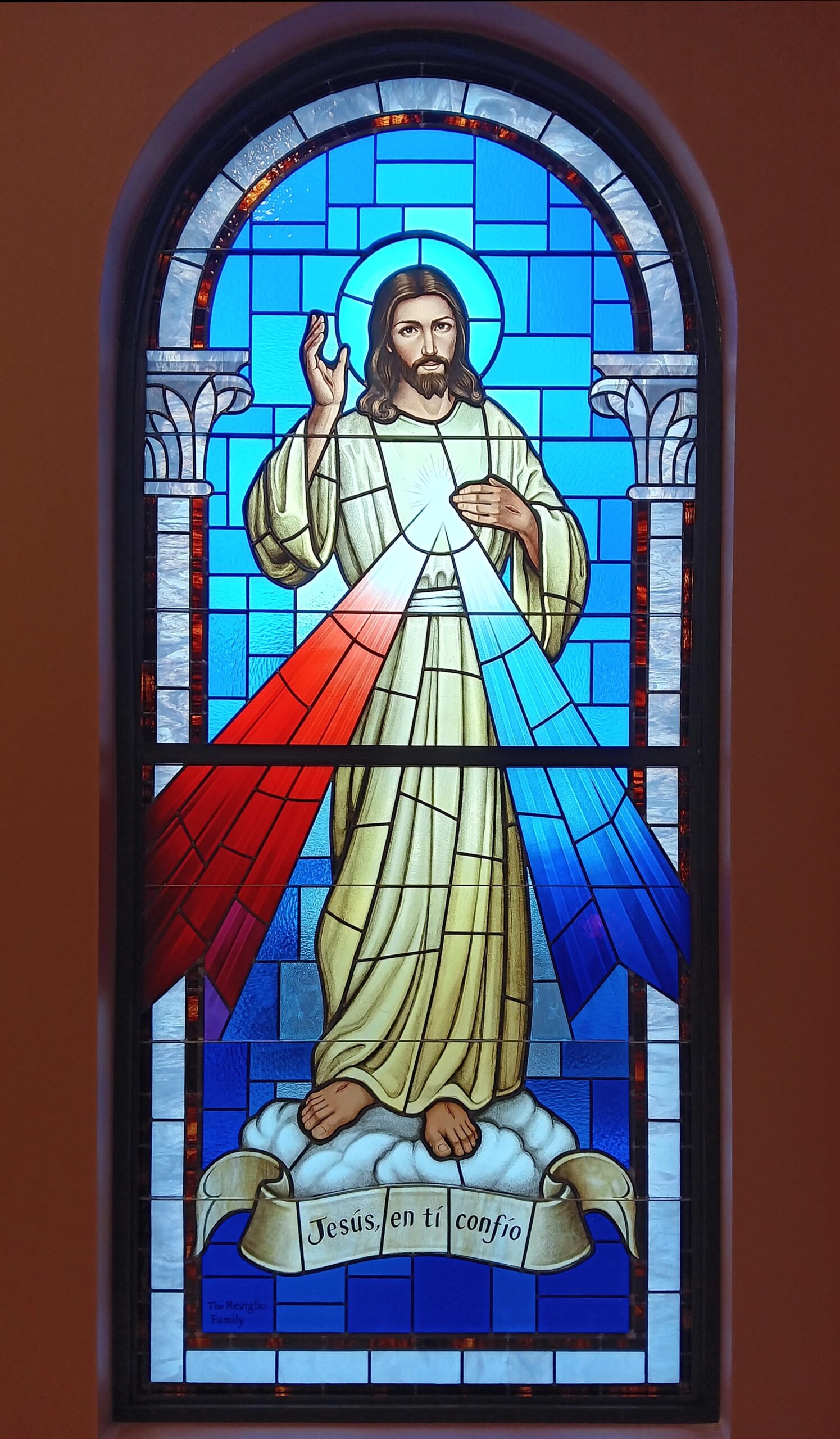 Stained glass window depicting Jesus with outstretched hand, surrounded by red and blue rays, featuring the inscription "Jesús, en ti confío," symbolizing faith and inspiration for worship spaces.