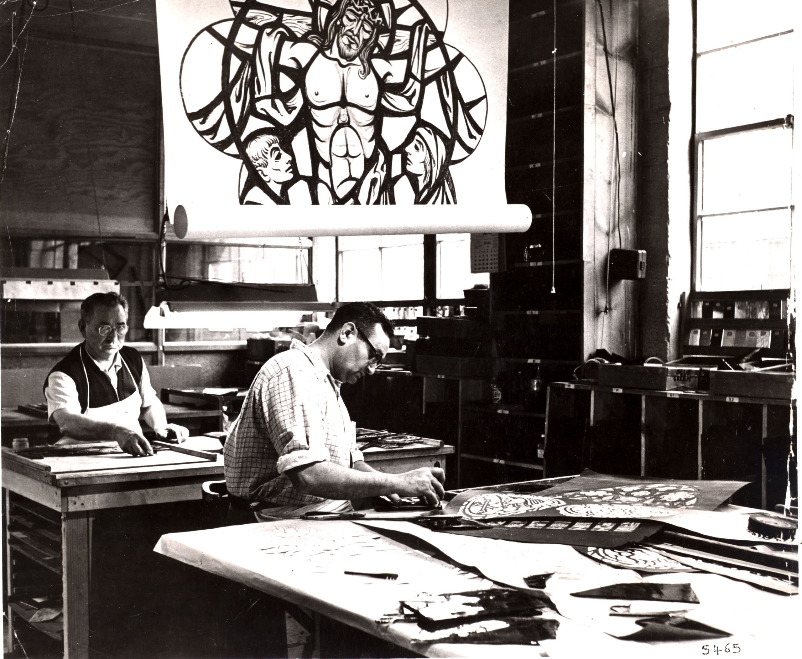 Two artisans working in a stained glass studio, one sketching designs and the other assembling glass pieces, with a large stained glass artwork featuring religious imagery in the background, reflecting the craftsmanship and artistry of Willet Stained Glass Studios.
