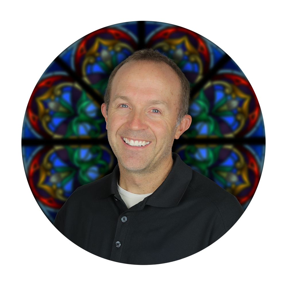 Greg Schow, partner at Willet Stained Glass Studios, smiling in front of a colorful stained glass background, representing the artistry and craftsmanship of the company.