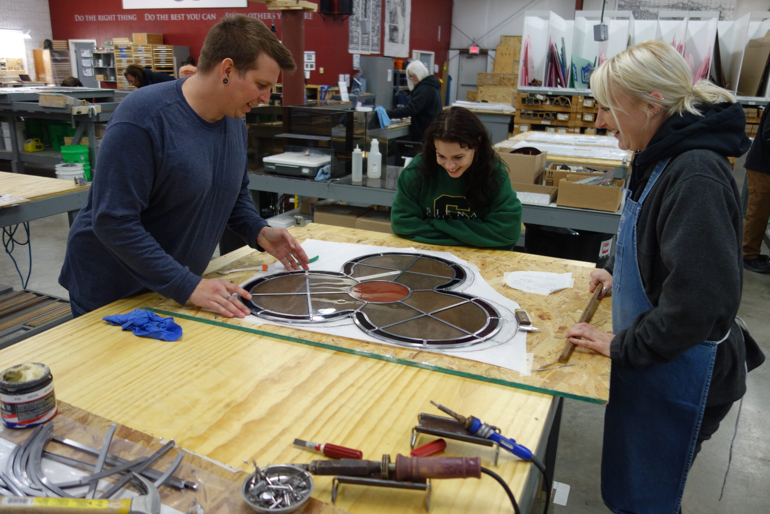 Three individuals collaborating on stained glass window design at Willet Studios, showcasing craftsmanship and teamwork in a workshop setting.