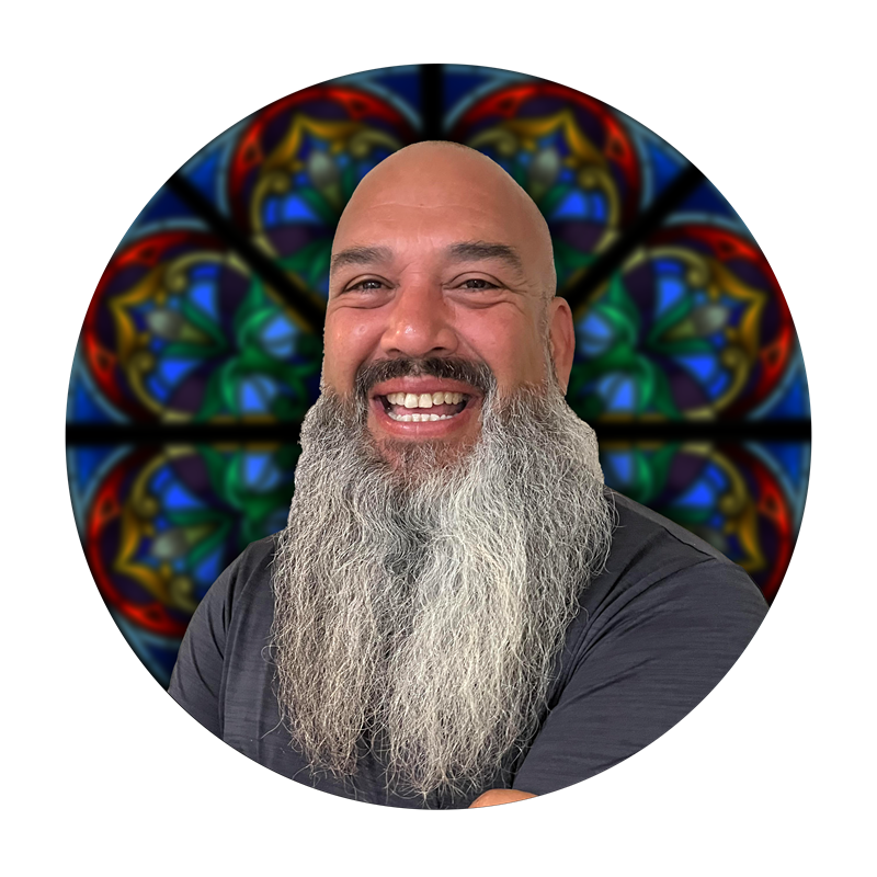 Smiling man with a beard in front of a colorful stained glass window, representing Willet Stained Glass Studios' craftsmanship and team spirit.