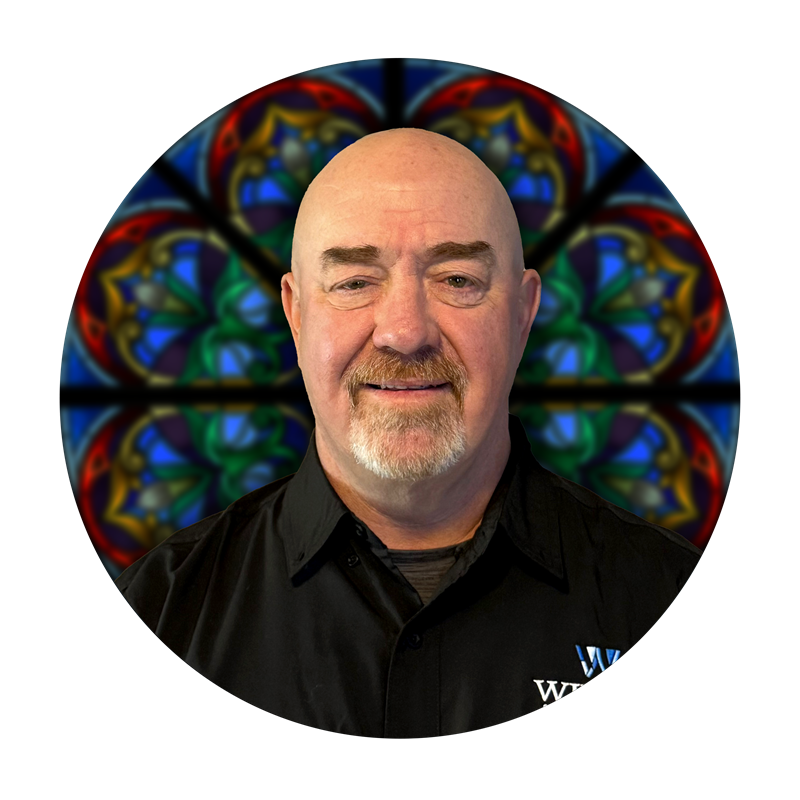 Tim Hudson, Sr. Studio Consultant at Willet Stained Glass Studios, smiling in front of a colorful stained glass background.