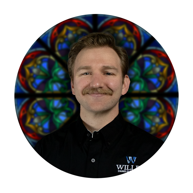 Zack Krings, Production Specialist at Willet Stained Glass Studios, smiling against a backdrop of colorful stained glass artistry.