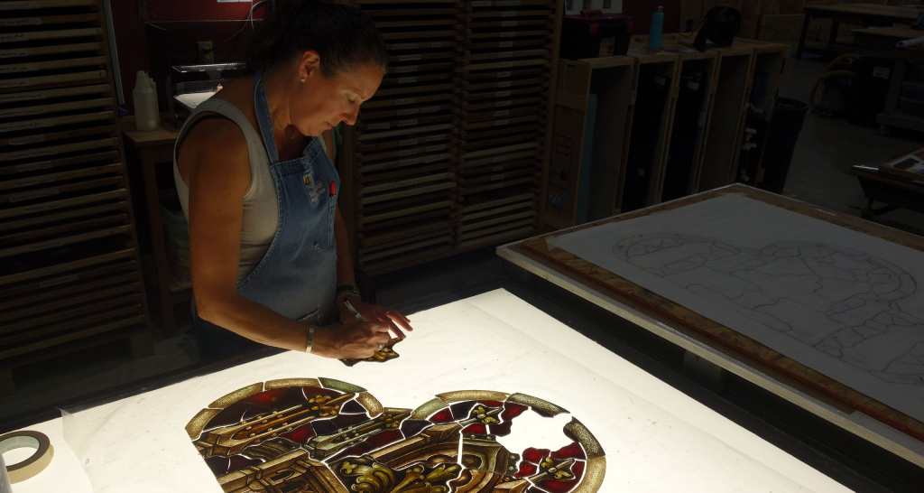 Woman restoring stained glass window in workshop, focusing on intricate details and craftsmanship, highlighting stained glass restoration process.
