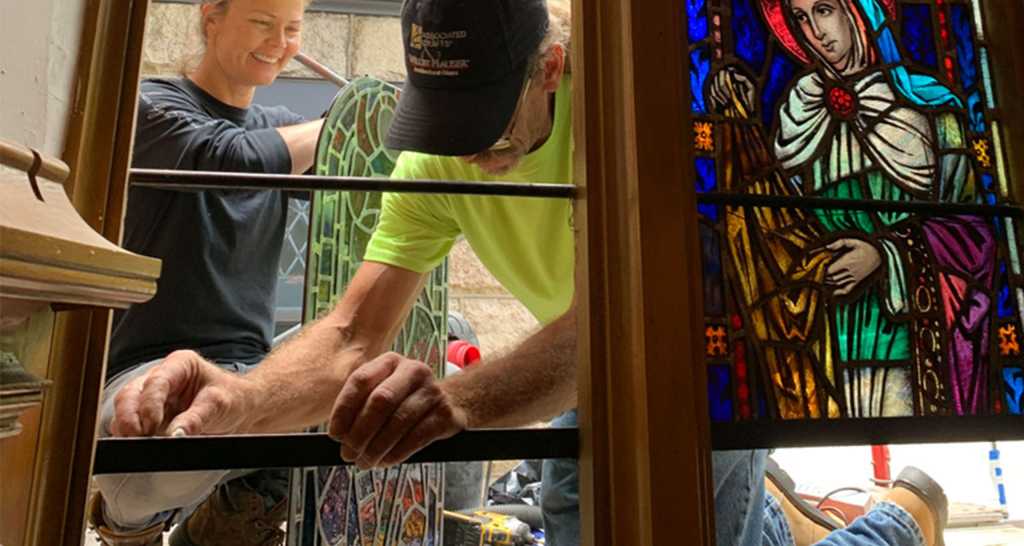 Workers installing stained glass window featuring a religious figure, showcasing the artistry and craftsmanship involved in stained glass repair and restoration.