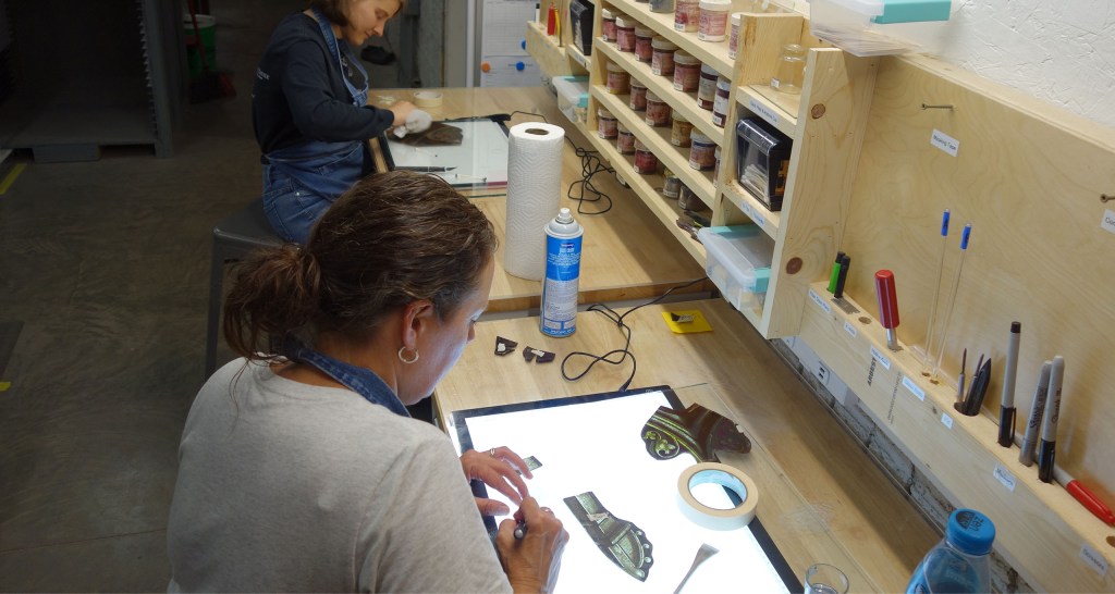 Two artisans working on stained glass restoration, focusing on intricate details, with tools and materials organized on a workbench in a studio setting.