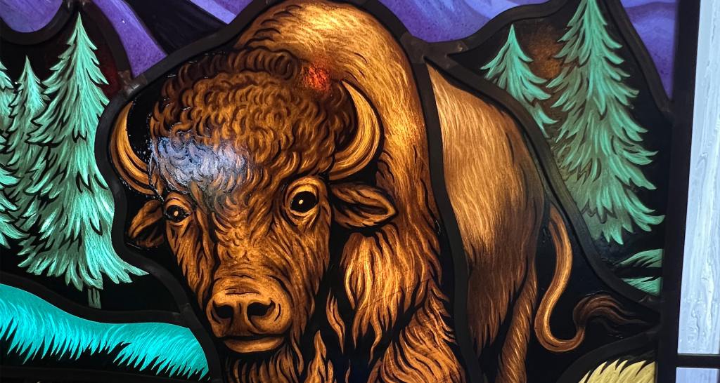 Bison depicted in stained glass artwork surrounded by trees and mountains, showcasing the beauty of architectural art glass in historical and spiritual contexts.