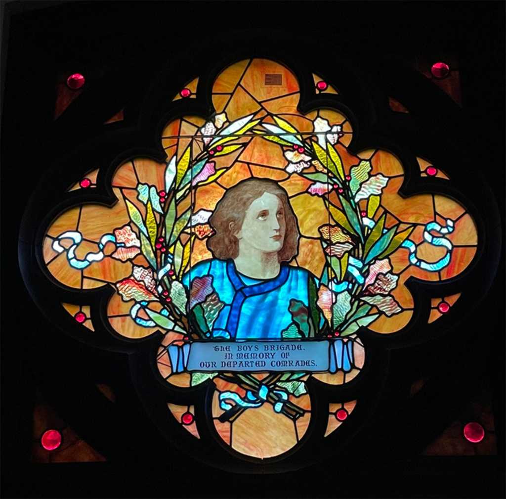 Stained glass window featuring a young figure in blue surrounded by floral elements, inscribed with "The Boys Brigade, In Memory Of Our Departed Comrades," symbolizing historical and spiritual narratives in church architecture.