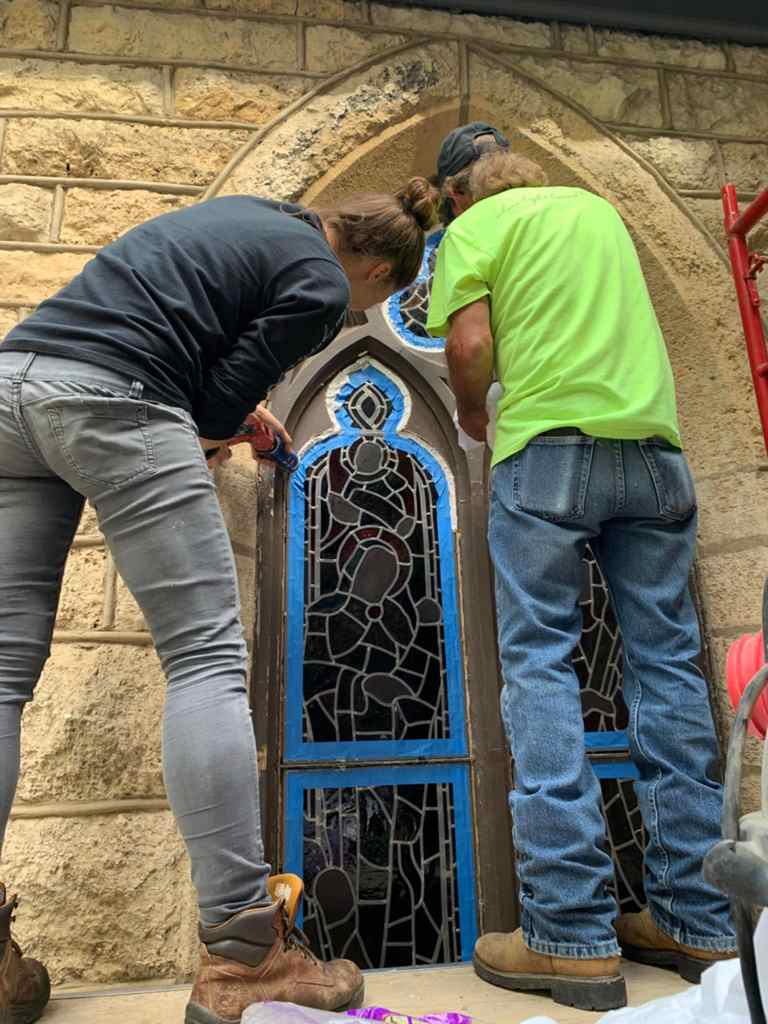 Workers restoring a stained glass window, showcasing craftsmanship and attention to historical detail, in a stone building.